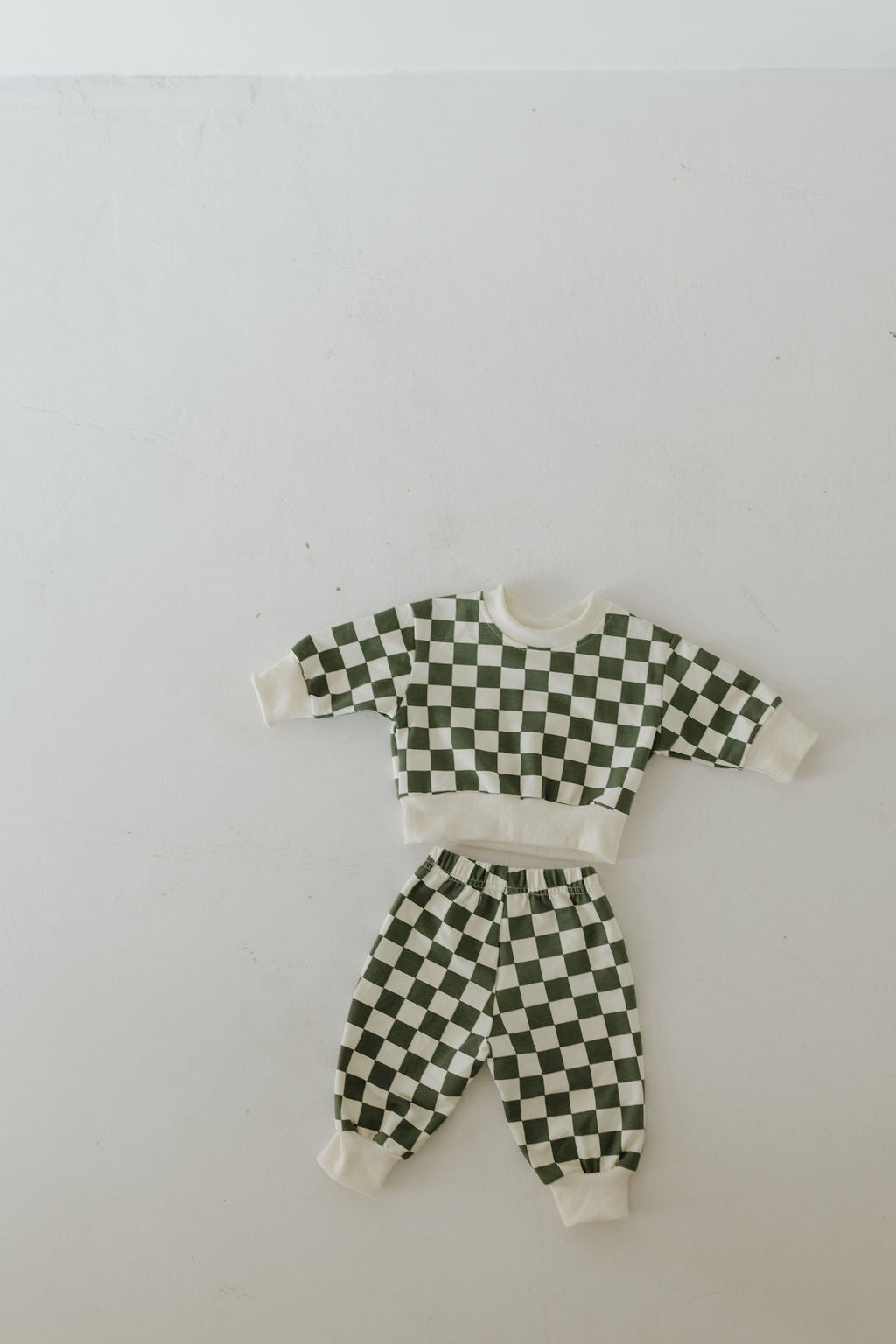 A small child's outfit, the "Sweat Set | Lucky" by forever french baby, is laid out flat on a white surface. It features a long-sleeve top and matching pants with green and white checkerboard patterns.