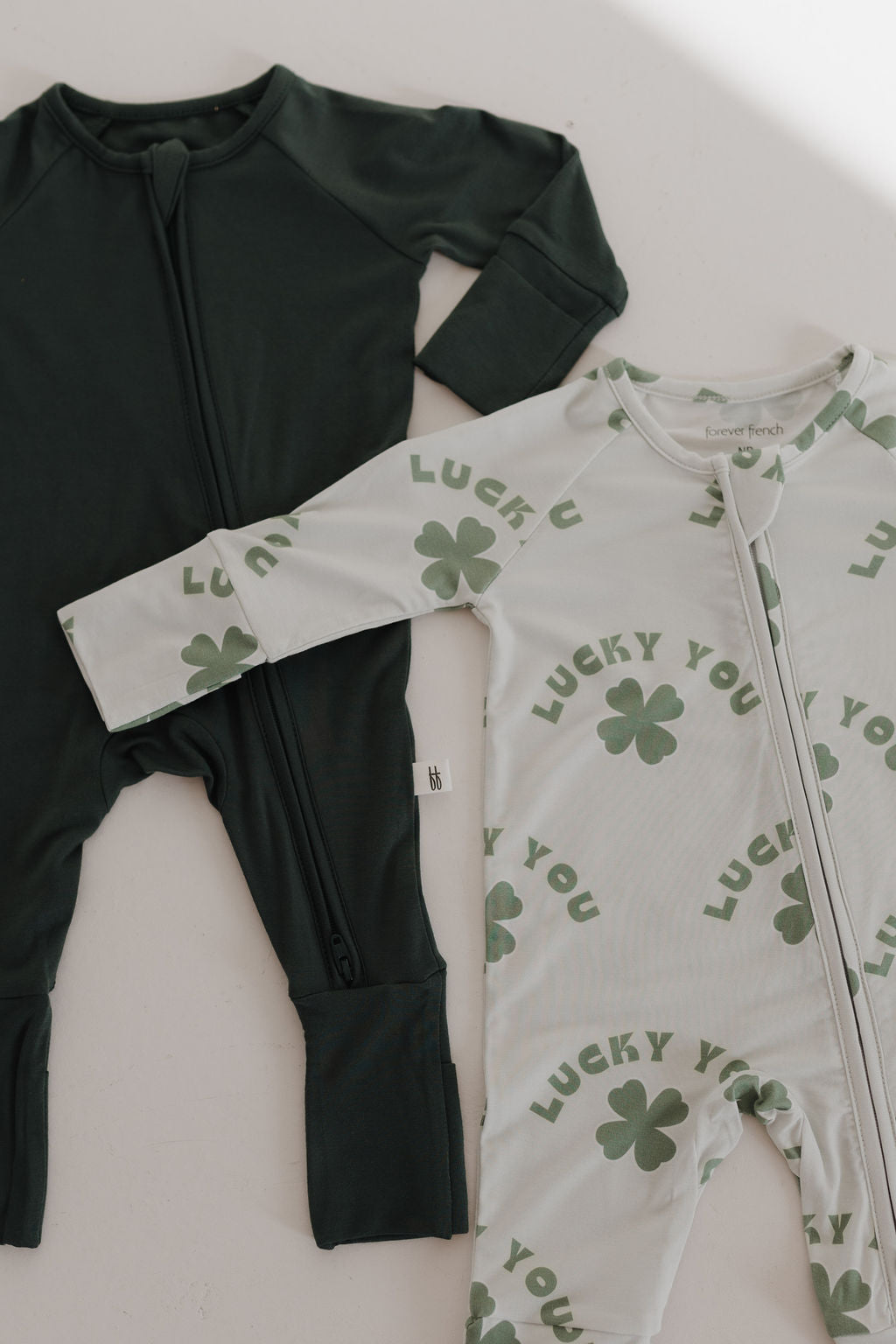 Two bamboo zip pajamas from the brand forever french baby are displayed. One is solid dark green, and the other is white with green clover prints and "Lucky You" text. Both feature front zippers for convenience.