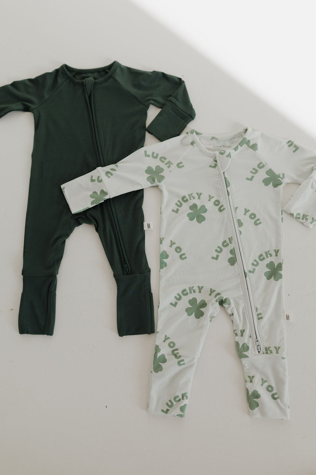 Two onesies from forever french baby are displayed: The Bamboo Zip Pajamas in solid dark green feature a breathable bamboo fabric and zipper front. The other is white with green shamrocks, "Lucky You" text, and an easy-zip design for hypo-allergenic comfort.