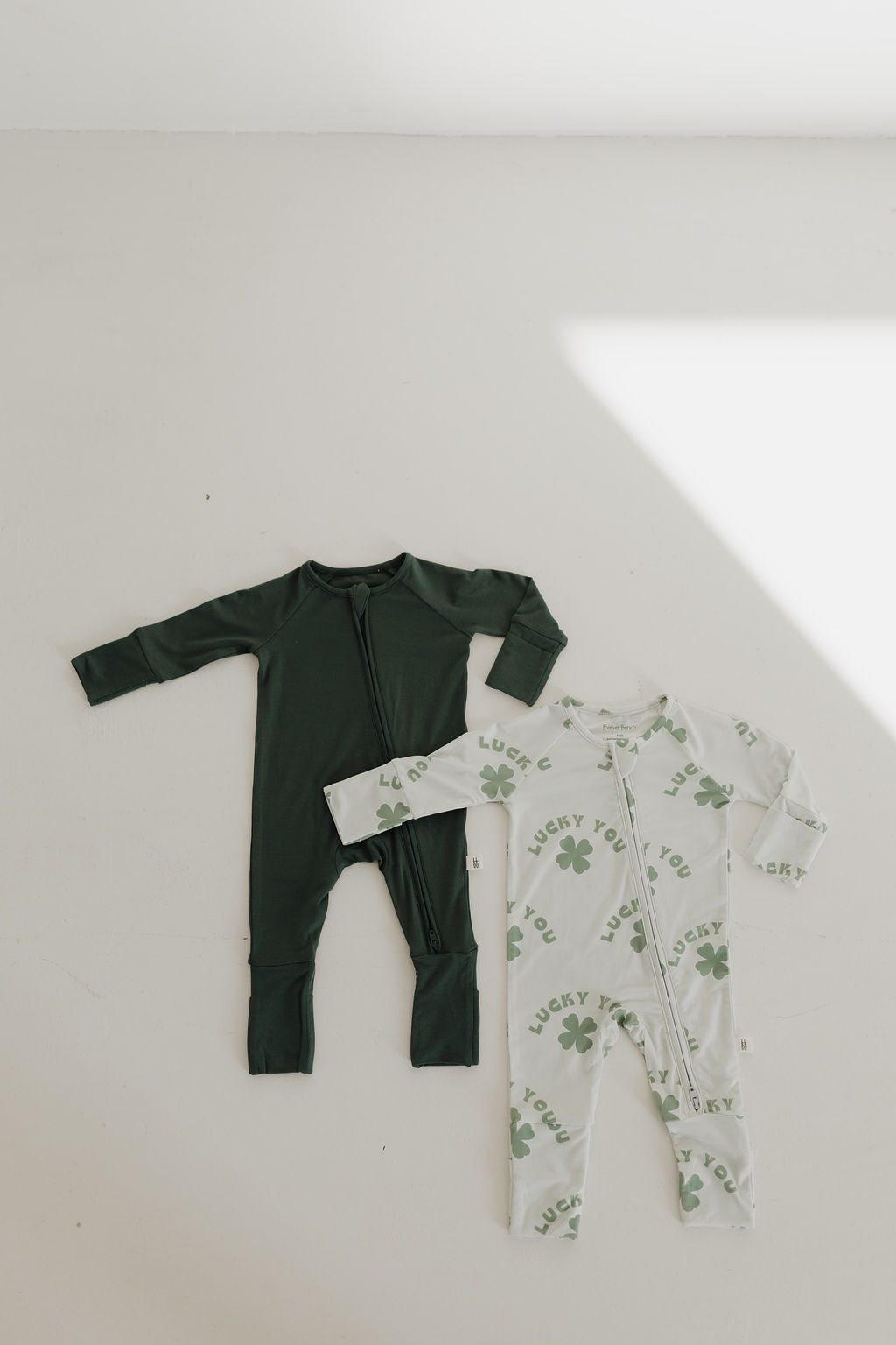 Two hypo-allergenic baby onesies are laid out on a light background—one is Bamboo Zip Pajamas in Emerald by forever french baby, and the other is white with green four-leaf clovers and "LUCKY YOU!" in a playful font. They are perfect for sensitive skin, casting a sharp shadow across the scene.