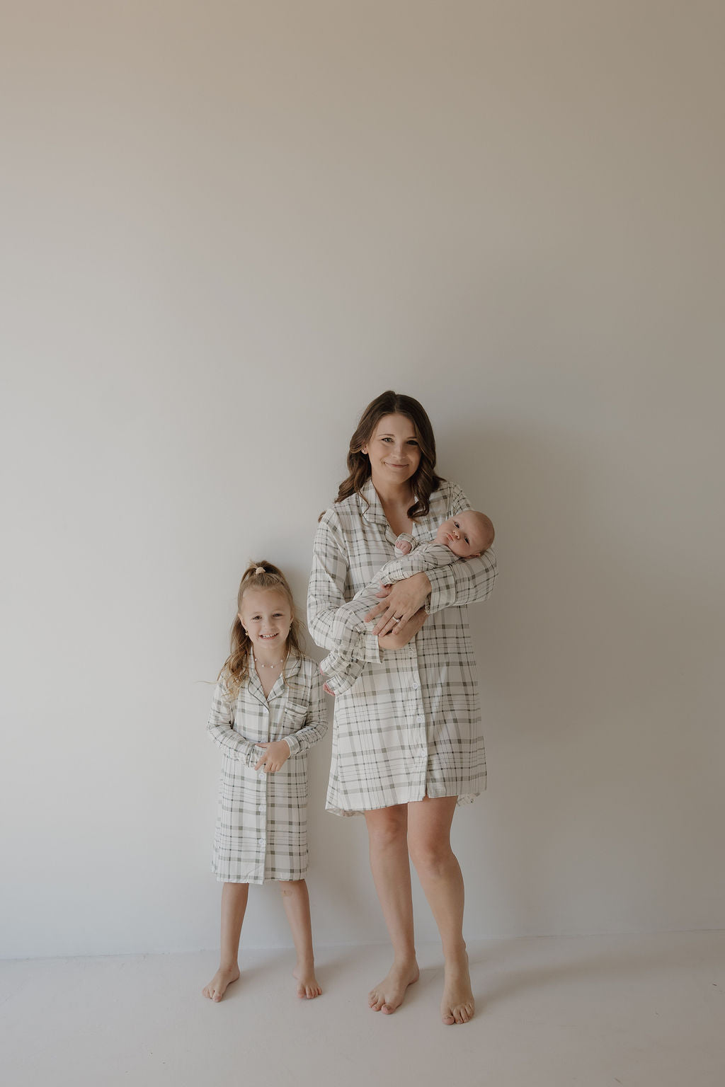 A woman with a baby and young girl stand barefoot by a plain wall, all smiling softly in matching forever french baby Picnic Plaid bamboo sleeping dresses.