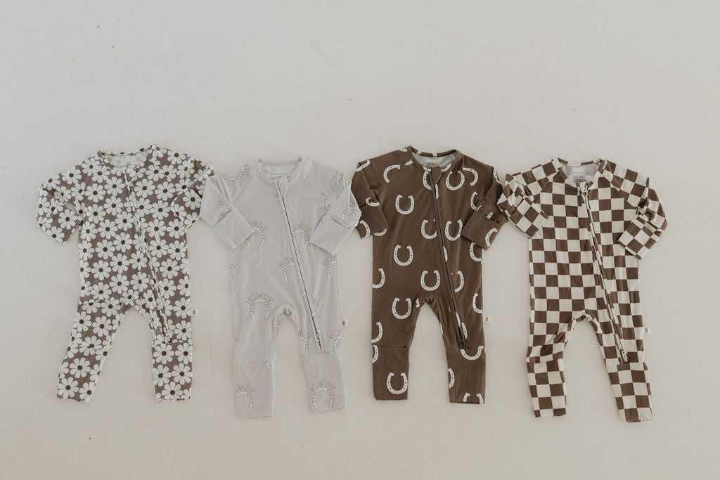 Displayed on a light surface are four Bamboo Zip Pajamas from forever french baby: a floral pattern in brown and white, a white onesie with soft line designs, a brown onesie with white horseshoes, and a brown and white checkered pattern. These offer the perfect cozy comfort.