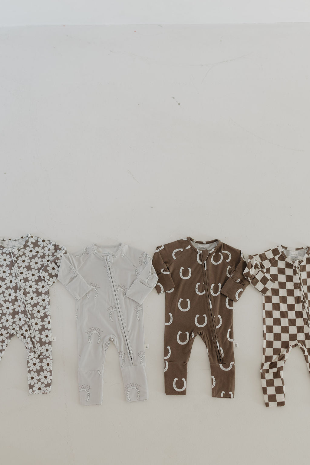 Four Bamboo Zip Pajamas from forever french baby are displayed on a light surface. They feature floral, pastel blue with rainbows, brown with horseshoes, and brown/beige checkered patterns. All are hypoallergenic to ensure comfort with breathable fabric for your little one's sensitive skin.