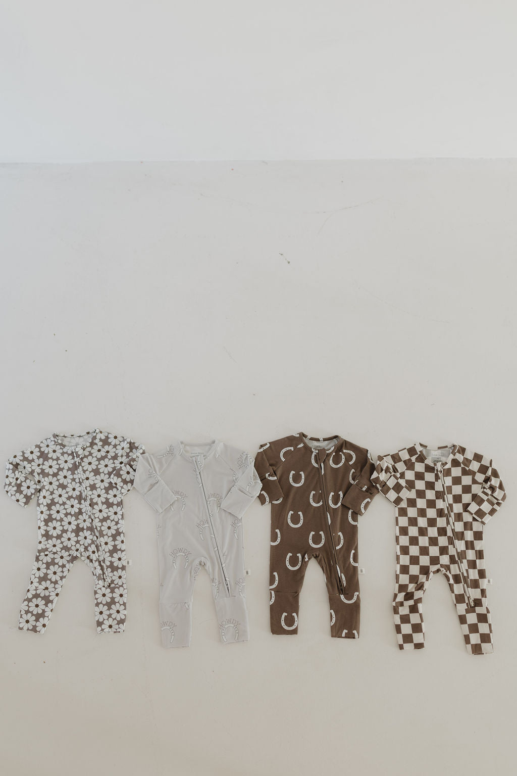 Four bamboo zip pajamas from forever french baby are displayed on a white surface, each with a unique pattern: white flowers on gray, plain gray, white moons on brown, and brown and white checkered. They're crafted from breathable fabric to keep your little one comfy all day.