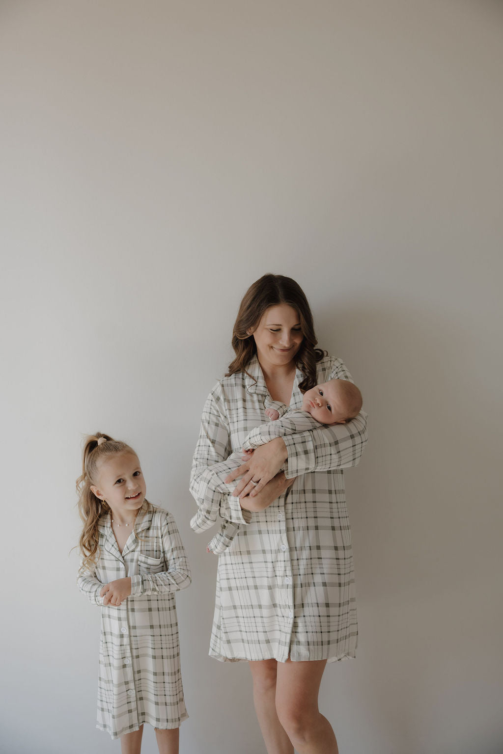A woman holds a baby as a young girl stands beside her; all three wear matching forever french baby Picnic Plaid bamboo sleeping dresses, posing against a plain, light-colored wall.