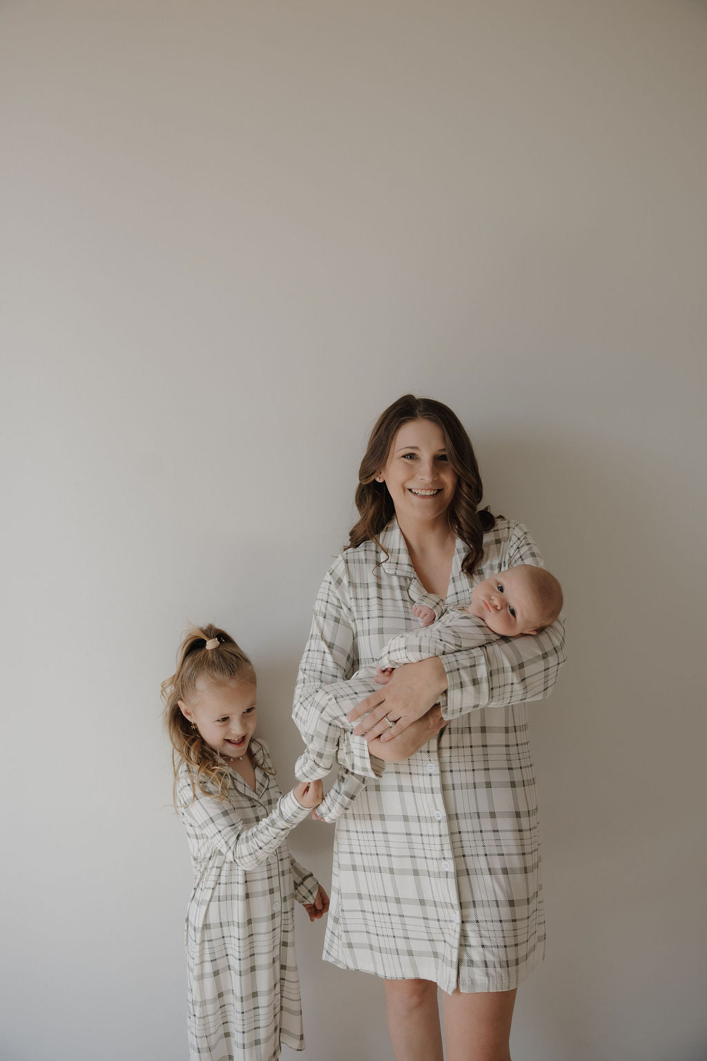A woman smiles holding a baby, with a young girl beside her reaching out. All three wear "forever french baby" Picnic Plaid outfits, including the Women's Bamboo Sleeping Dress, made from soft, breathable fabric against a light-colored wall.