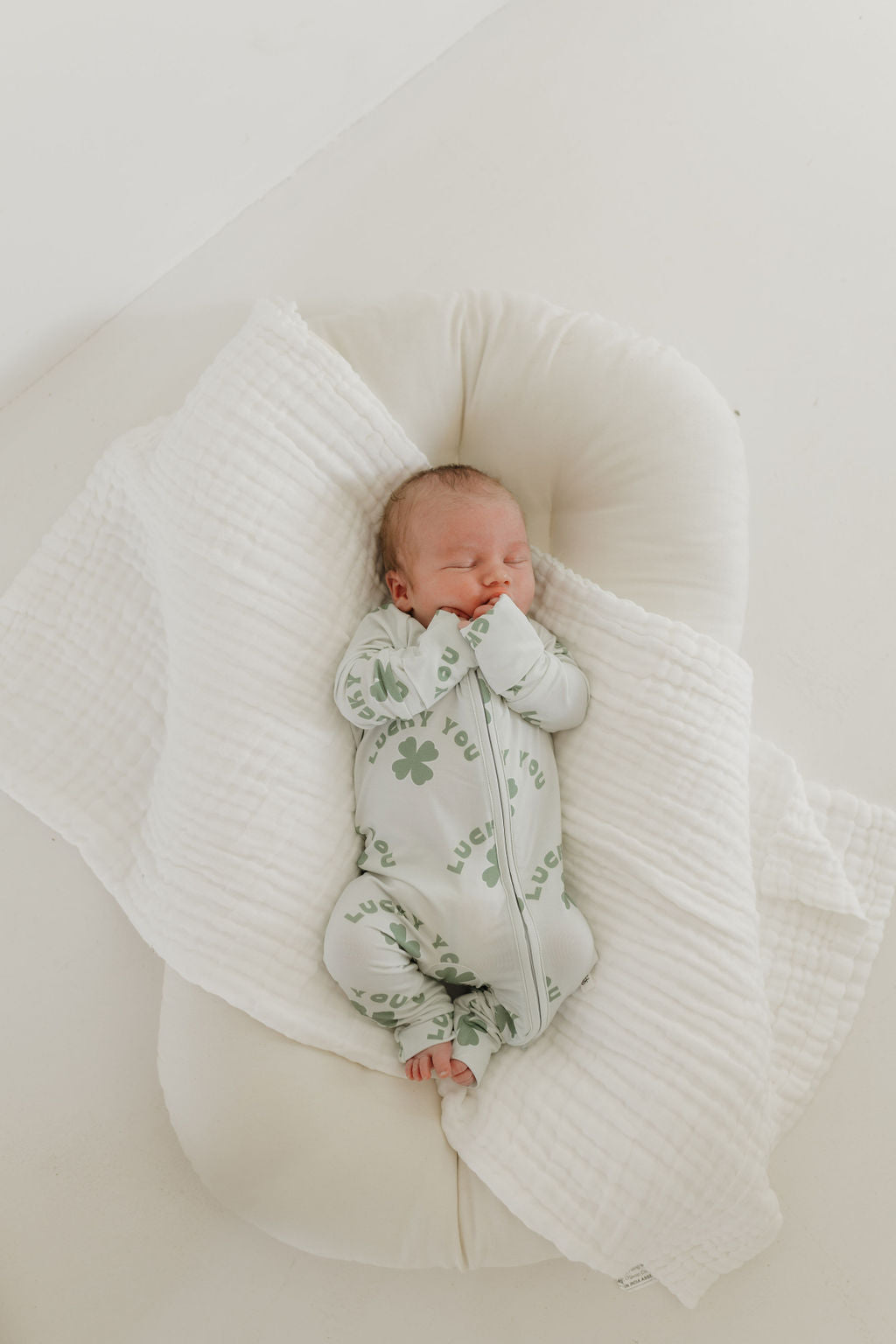 A baby is napping on a white cushioned surface, partially covered with a soft white blanket. The baby wears "Bamboo Zip Pajamas | Lucky You," light green onesie by forever french baby, adorned with shamrocks and the word "Lucky," made from breathable fabric ideal for sensitive skin.