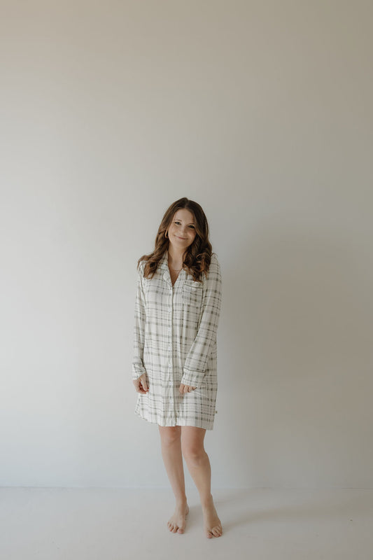 A woman with long brown hair stands barefoot against a plain wall, smiling in the forever french baby Women's Bamboo Sleeping Dress | Picnic Plaid—crafted from breathable, gentle, hypo-allergenic fabric.