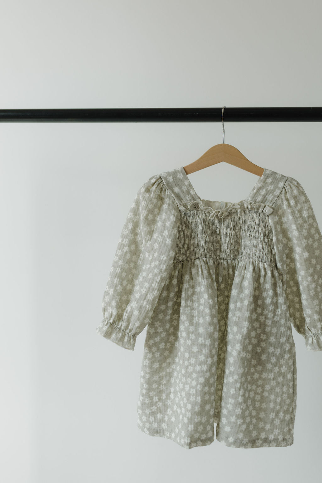 The hypo-allergenic, floral-patterned Child Dress in French Gray by forever french baby hangs elegantly on a wooden hanger against a plain white background. It features smocked detailing on the chest and is displayed on a black clothing rod.