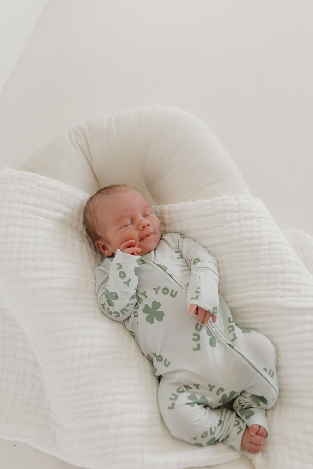 A peaceful scene shows a sleeping baby on a soft, white cushion, wrapped in forever french baby's hypoallergenic Bamboo Zip Pajamas. The light-colored onesie is adorned with green shamrocks and the phrase "Lucky You.