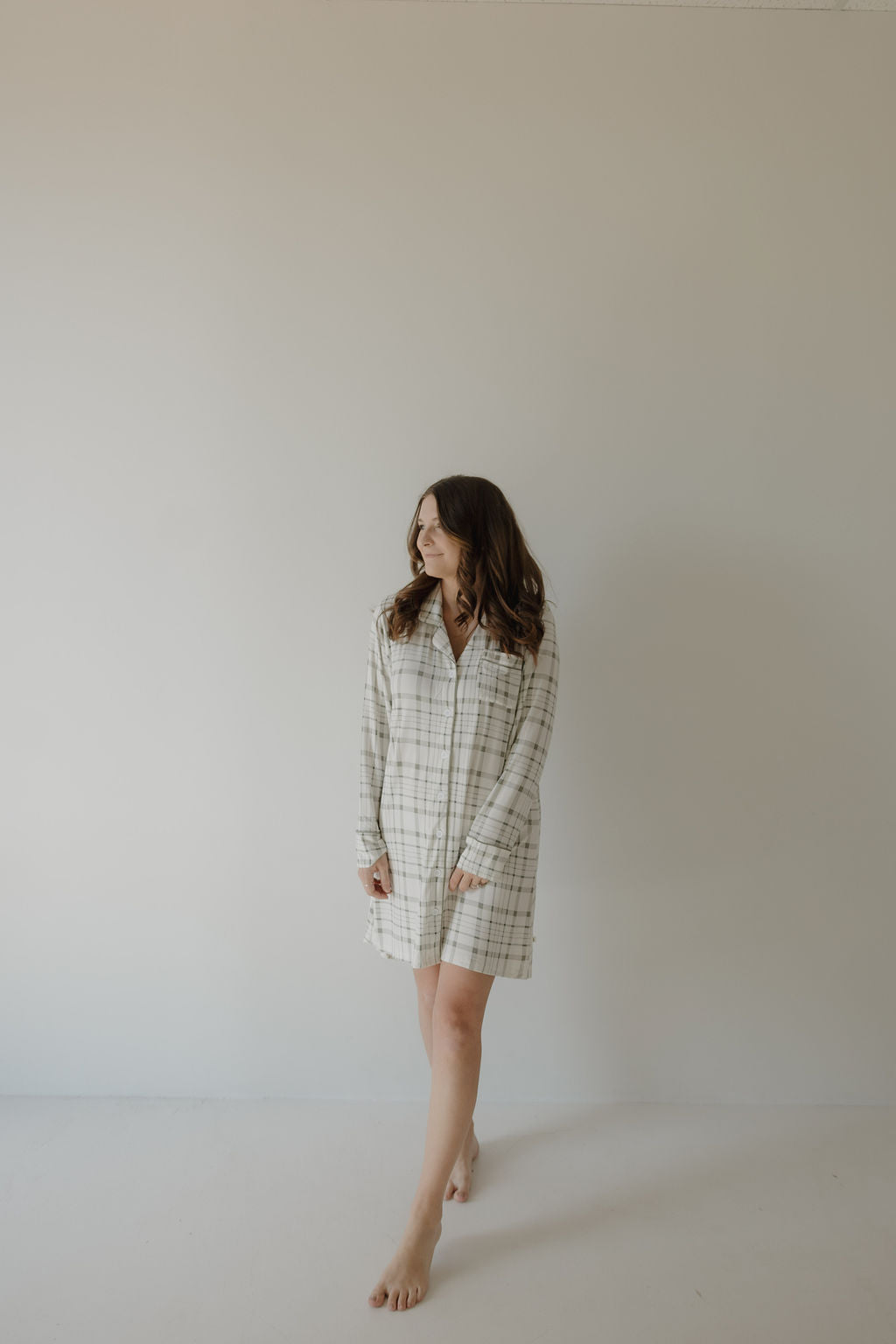 A woman with long brown hair stands barefoot by a light wall, wearing the forever french baby Women's Bamboo Sleeping Dress in Picnic Plaid. The loose-fit, breathable dress gives her a relaxed look as she gazes to the side.