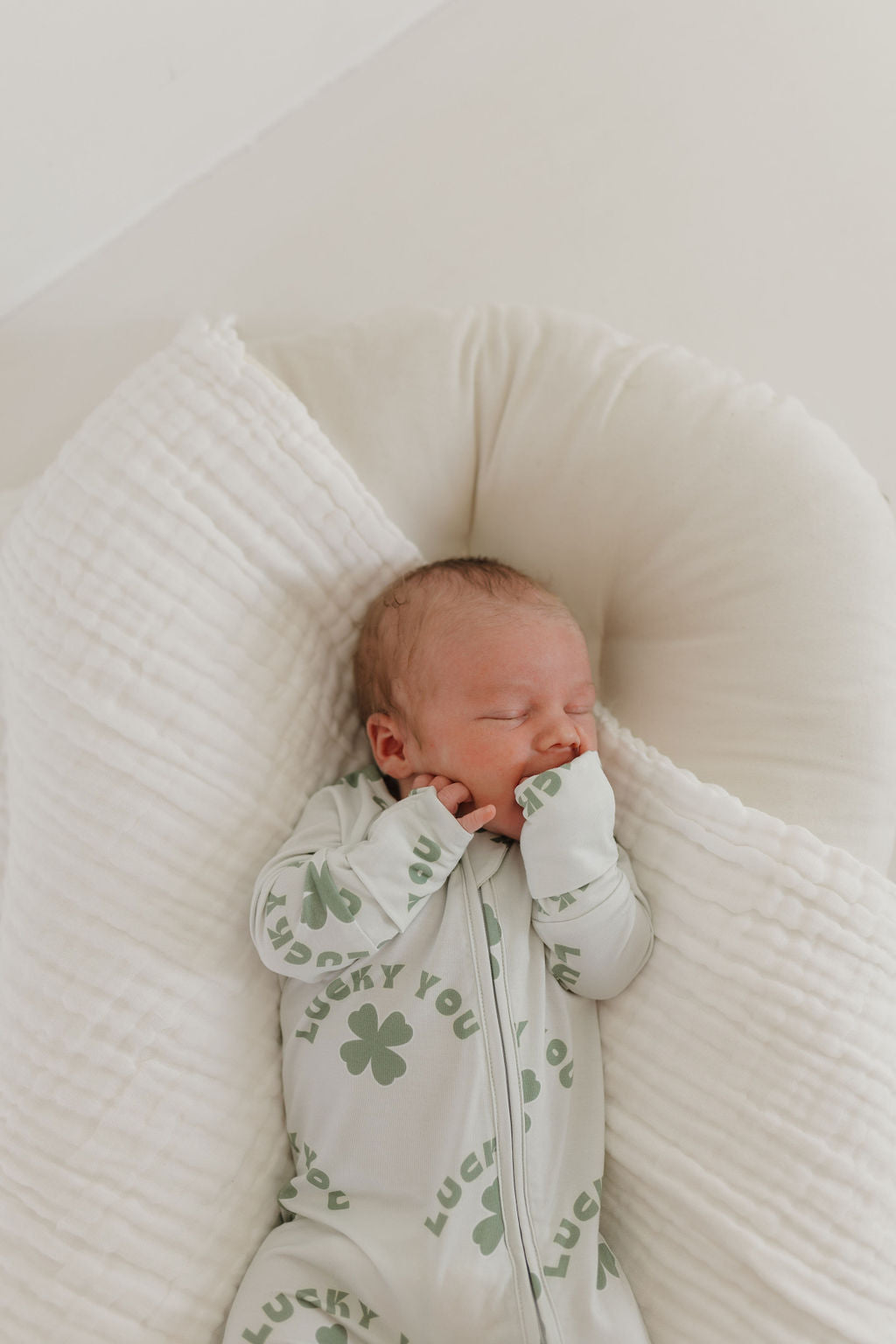 A newborn sleeps peacefully on a soft cushion, wrapped in breathable fabric, wearing forever french baby's "Bamboo Zip Pajamas | Lucky You," featuring a light green design with four-leaf clovers and the words "Lucky You." The baby's tiny hands rest near its face.