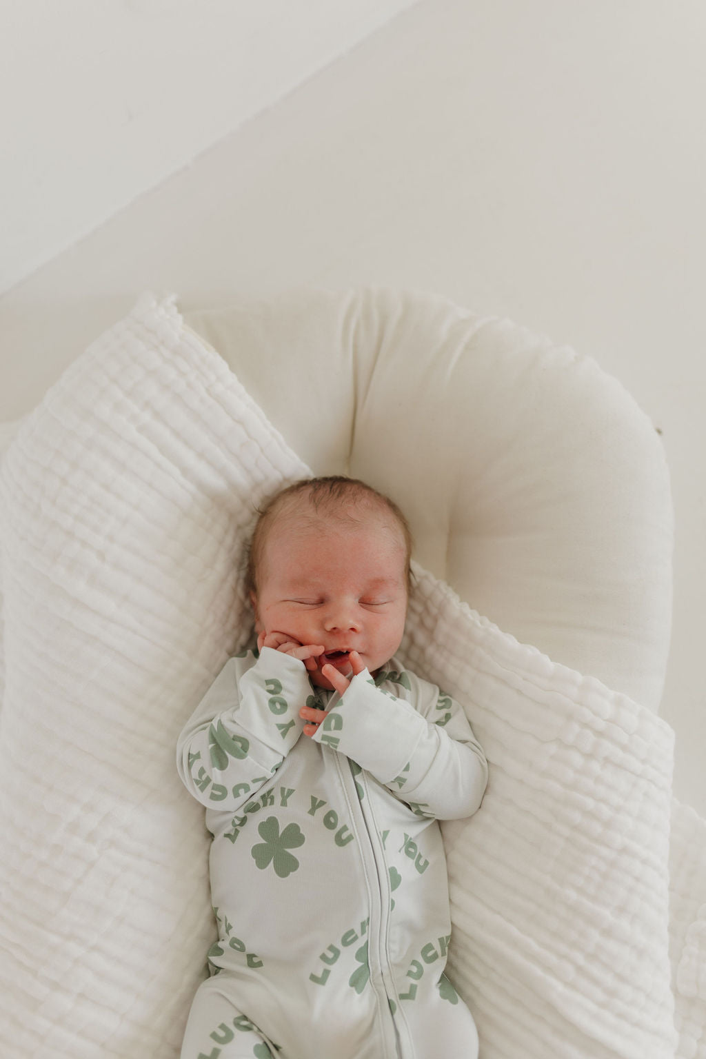 A newborn baby sleeps peacefully in a soft, white, cushioned nest, wearing Forever French Baby's "Lucky You" Bamboo Zip Pajamas. The cozy long-sleeve onesie features green clovers and a breathable fabric, creating a serene scene.