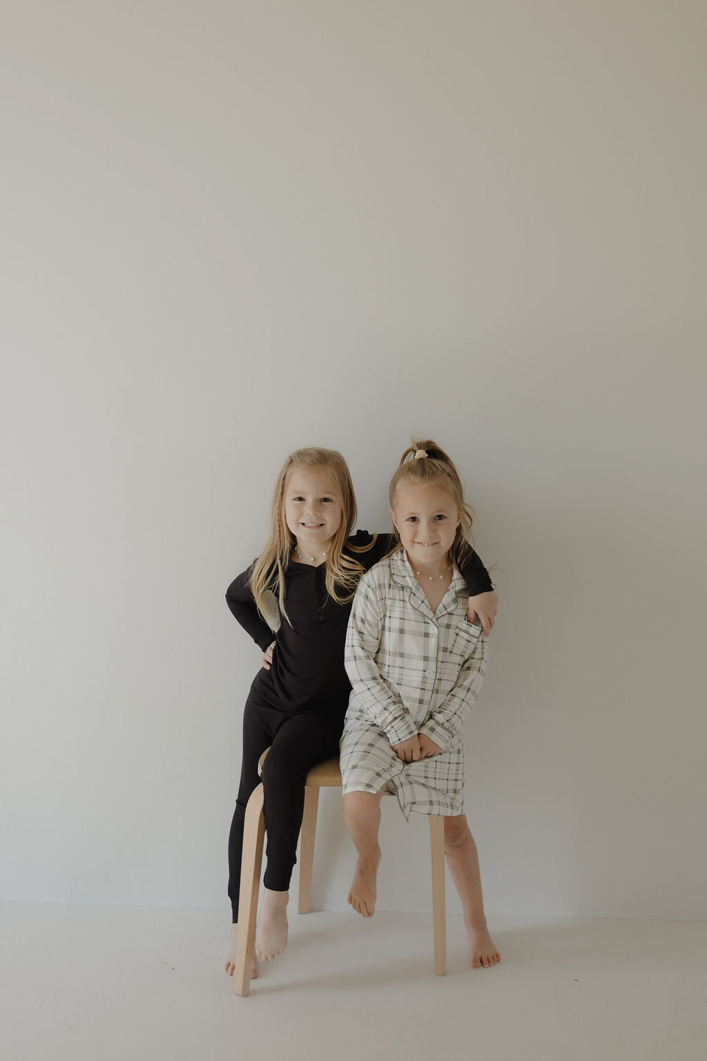 Two young girls pose in a minimal setting. One sits on a stool dressed in black, while the other smiles barefoot in the forever french baby Toddler Bamboo Sleeping Dress | Picnic Plaid, made from soft bamboo fabric.