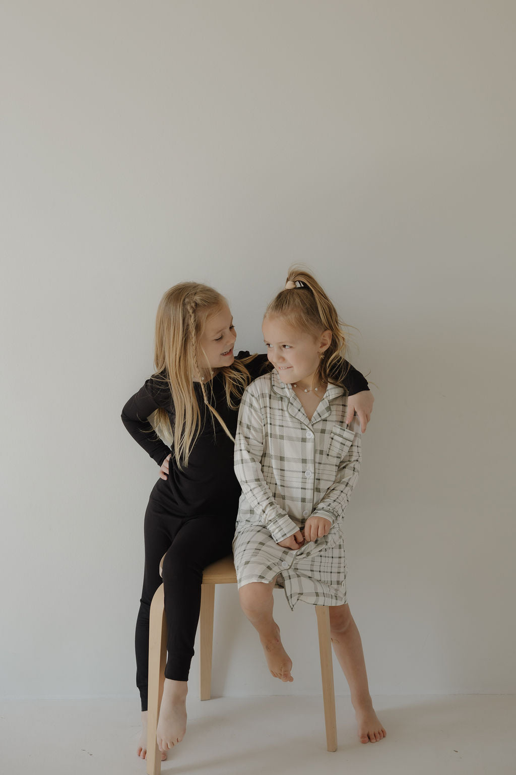 Two young girls sit on a wooden stool by a plain light wall. One wears the forever french baby Toddler Bamboo Sleeping Dress in Picnic Plaid, while the other sports hypo-allergenic sleepwear. They smile warmly at each other.