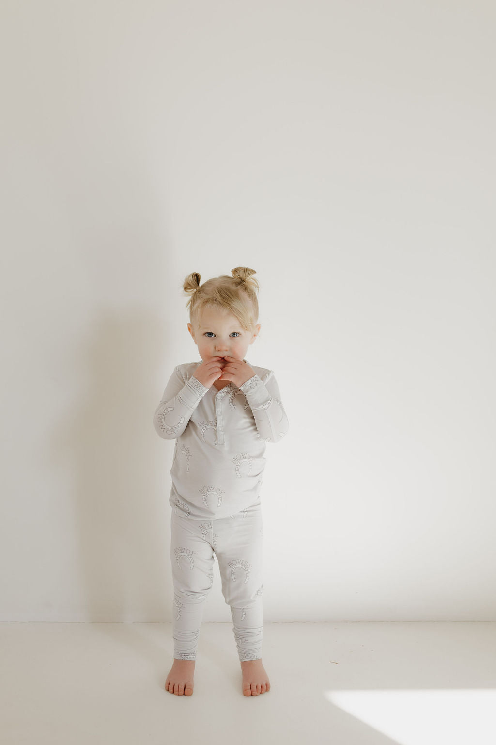 A toddler, featuring two small buns in their hair, stands against a plain white backdrop wearing forever french baby Bamboo Two Piece Pajamas | Howdy. The breathable fabric and subtle patterns of the pajamas match their curious expression as they gaze at the camera.