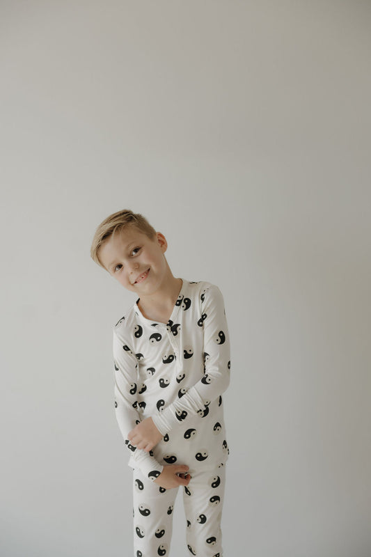 A young blond boy smiles in forever French baby's Bamboo Two Piece Pajamas | Black & White Yin Yang, made with breathable, hypo-allergenic fabric for comfort against a plain light background.
