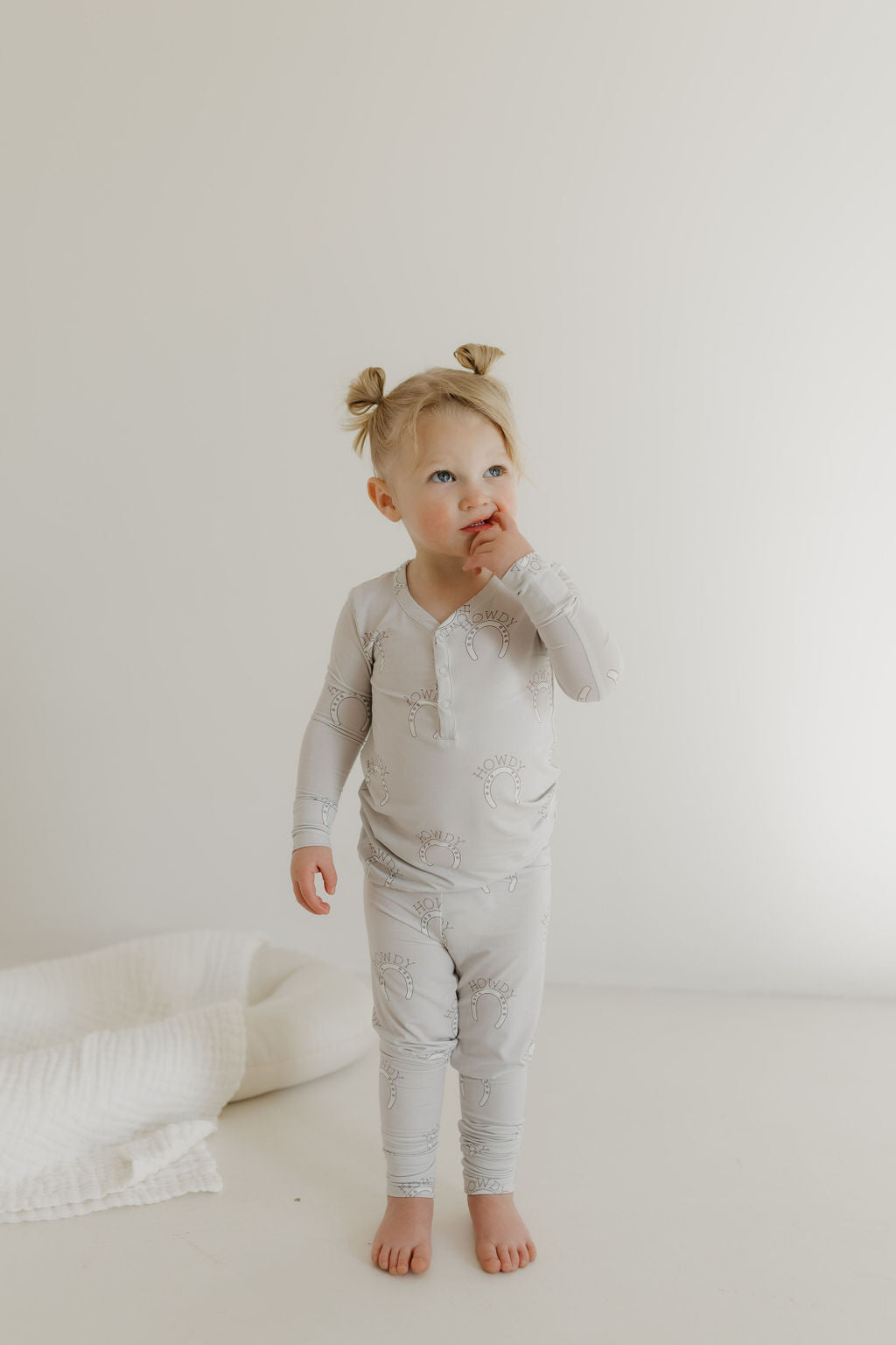 A toddler with light hair in pigtails stands barefoot on a light-colored floor, wearing the Bamboo Two Piece Pajamas by forever french baby. The cozy gray set is breathable, and they gaze upward with a finger to their lips. A folded white blanket nearby adds to the serene scene.