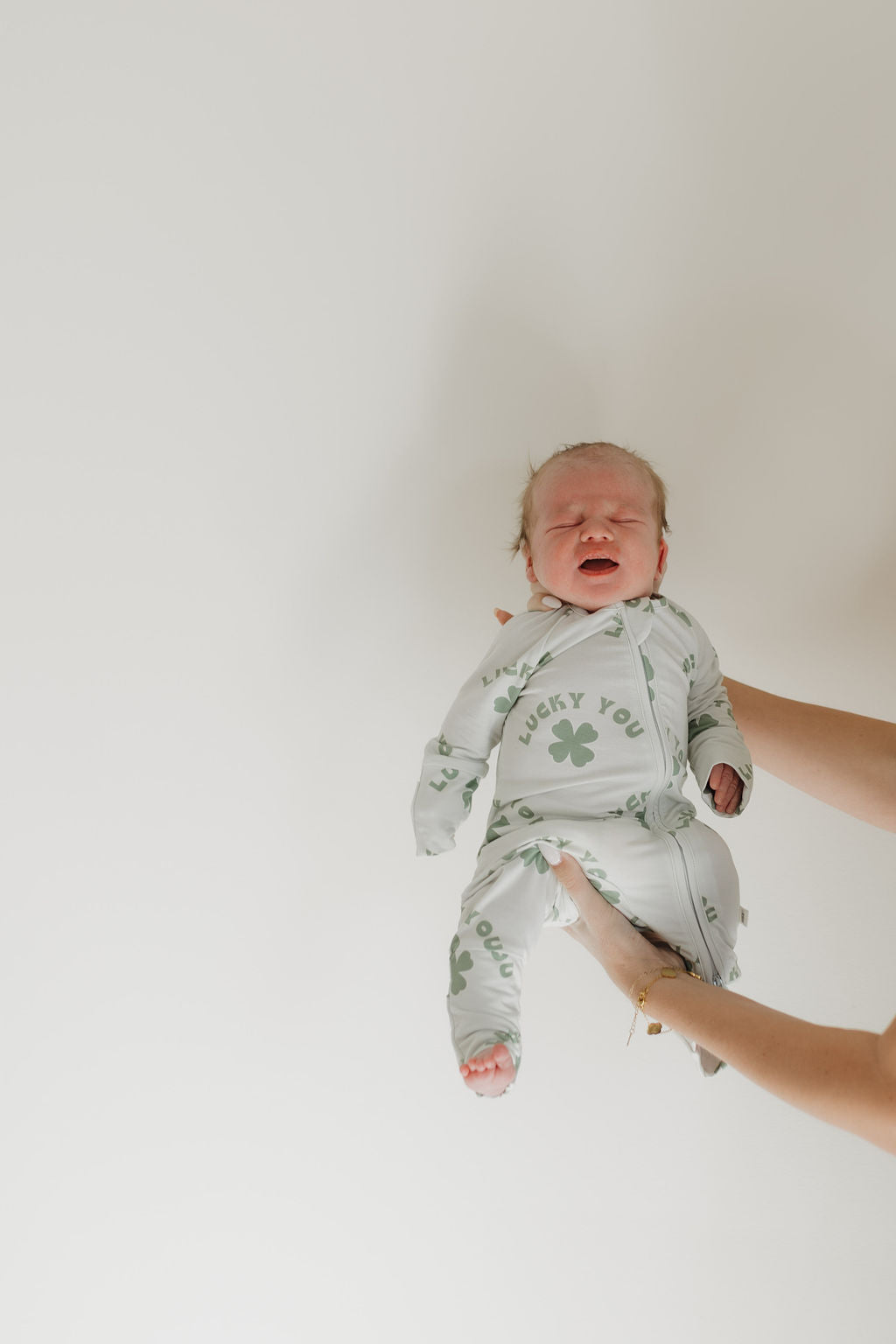 A calm baby wearing forever french baby's Bamboo Zip Pajamas in pale green, printed with "Lucky You," is gently held by two adult hands against a plain white background. The baby has closed eyes and a slight smile.