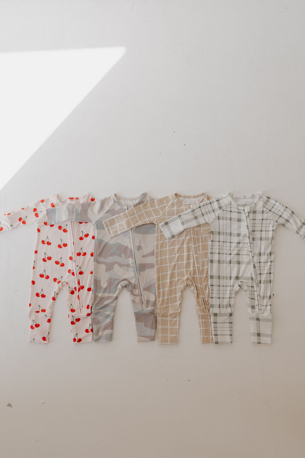 A set of four baby onesies is displayed on a light background. They feature cherry prints, camouflage, a beige grid design named "Bamboo Zip Pajamas | Grid" from forever french baby, and a plaid pattern. Made from breathable sleepwear material, these are hypo-allergenic for all-day comfort.