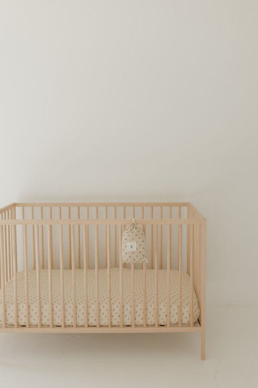 A minimalist wooden baby crib with the Forever French Baby Muslin Crib Sheet | Flora (Mis-Print Peach Tone) and a matching pillow, set against a plain white wall in a bright, simple room.