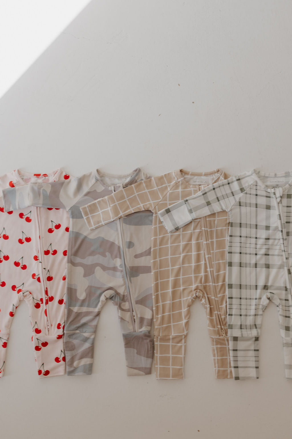 Four Bamboo Zip Pajamas by forever french baby are displayed side by side, featuring patterns like red cherries on white, gray camouflage, beige checkered, and gray plaid. These breathable onesies offer comfort for babies. Sunlight casts a diagonal shadow across the image.