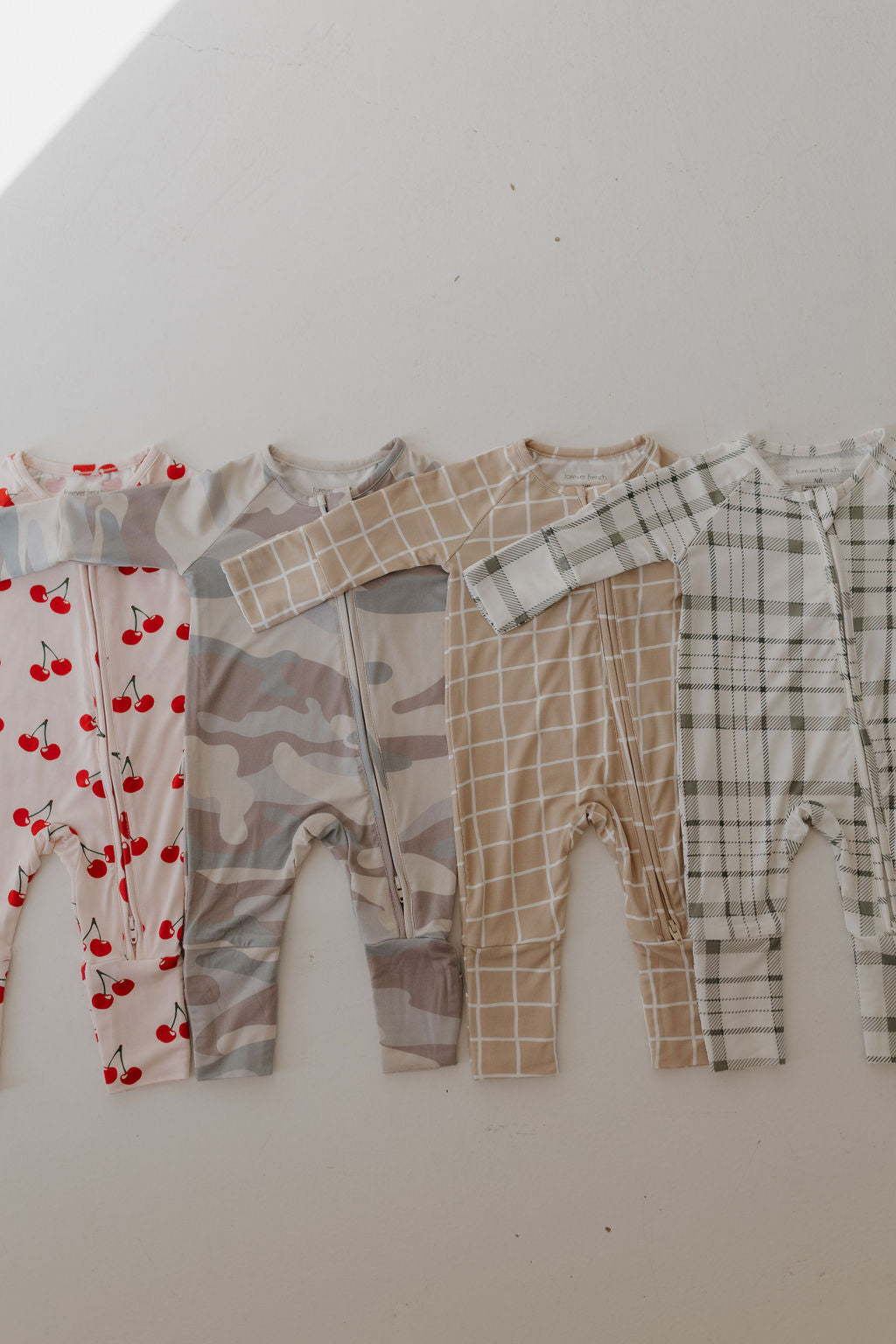 Four forever french baby Bamboo Zip Pajamas are displayed, showcasing delightful designs: cherries, camouflage, beige grid, and the Picnic Plaid. Made from soft fabric with long sleeves, they're breathable and ensure comfort for babies all day.