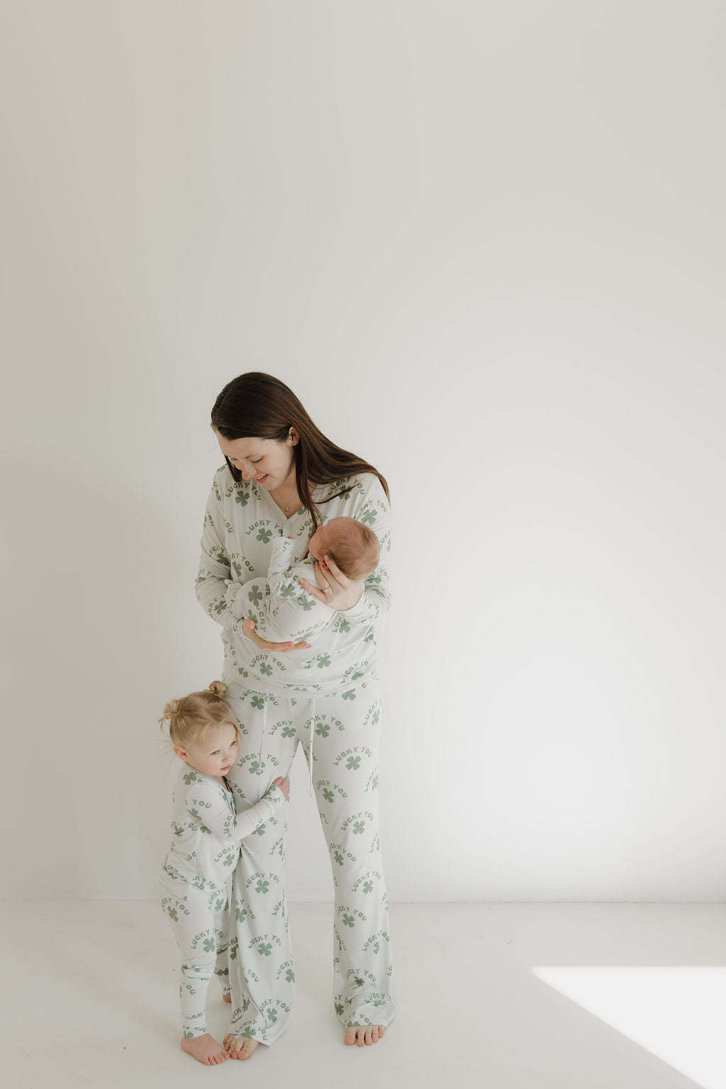 A woman stands against a white background, holding a baby as a young child clings to her leg. All wear matching "Lucky You" Bamboo Zip Pajamas from forever french baby, featuring a green leaf pattern with breathable fabric. Soft lighting highlights the comfort and elegance of their hypoallergenic attire.