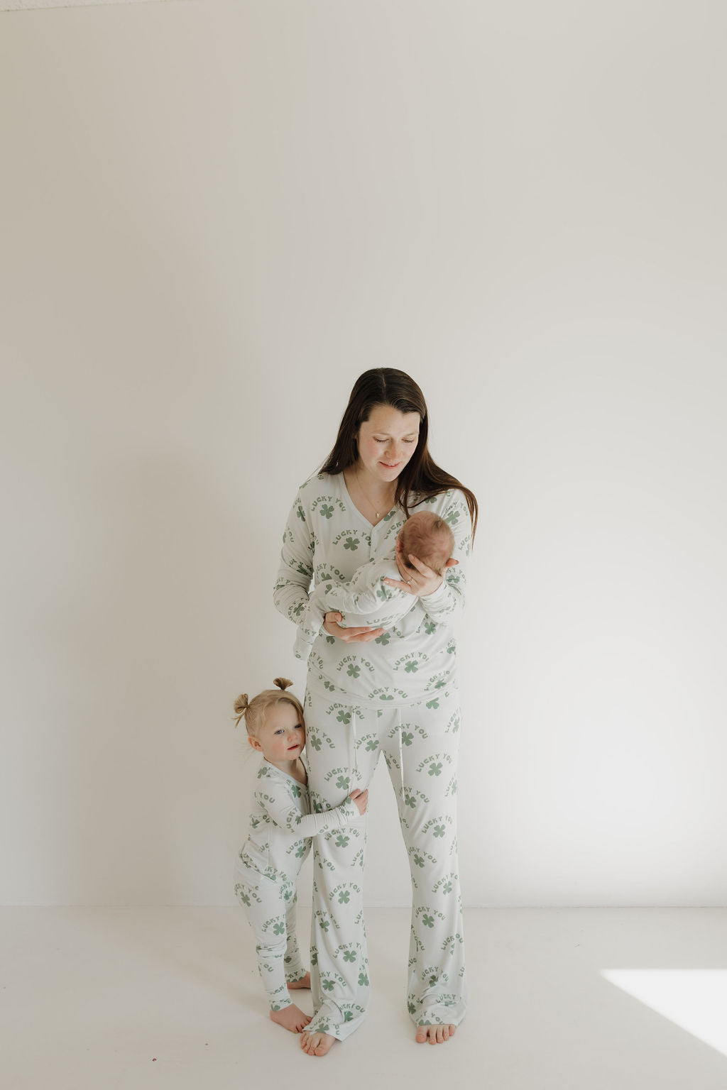 A woman wearing forever french baby's Women's Bamboo Pajama | Lucky You, with a pattern of green leaves on a light background, stands holding a baby while a young child hugs her leg in a softly lit, plain room with light-colored walls and floor.