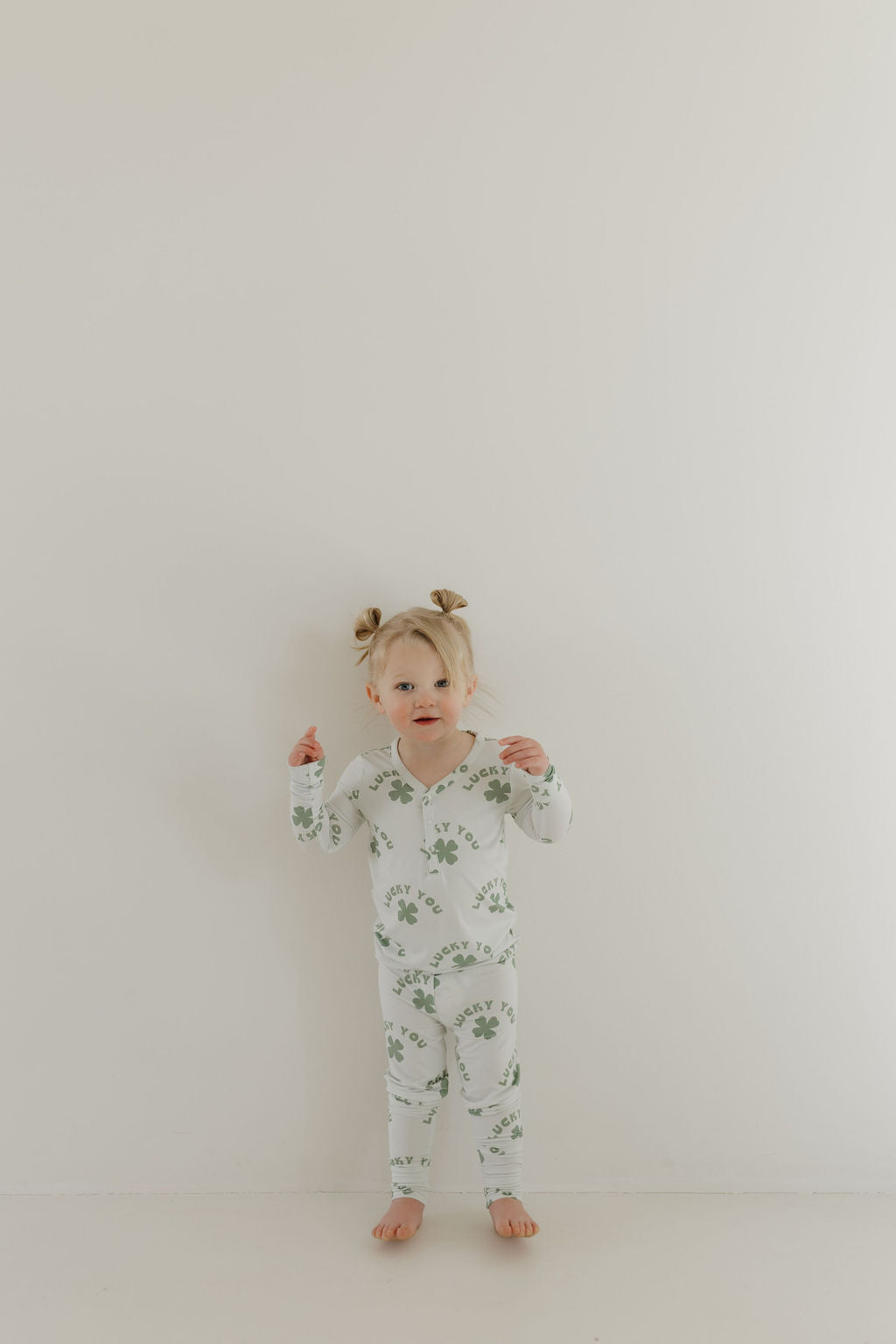 A young child with two small ponytails stands against a white wall, wearing the "Bamboo Two Piece Pajamas | Lucky You" by forever french baby. The light green, long-sleeve pajama set features a plant print and is made from breathable bamboo fabric. The child is barefoot and looking ahead neutrally.