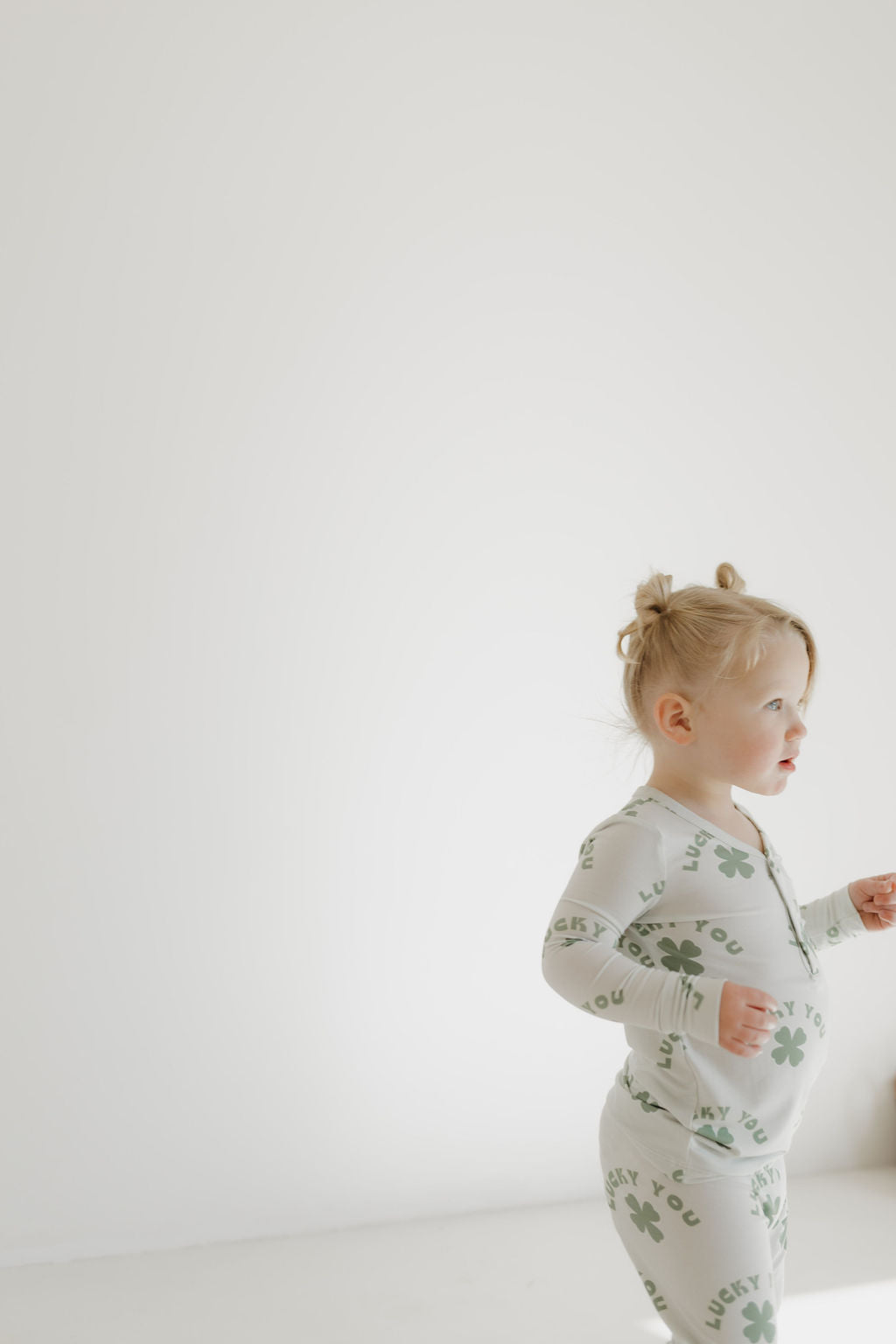 A toddler with blonde hair in small buns wears "Bamboo Two Piece Pajamas | Lucky You" by forever french baby, featuring green shamrocks and "LUCKY." Made from breathable, hypo-allergenic bamboo fabric for comfort, they walk to the right against a plain white background.