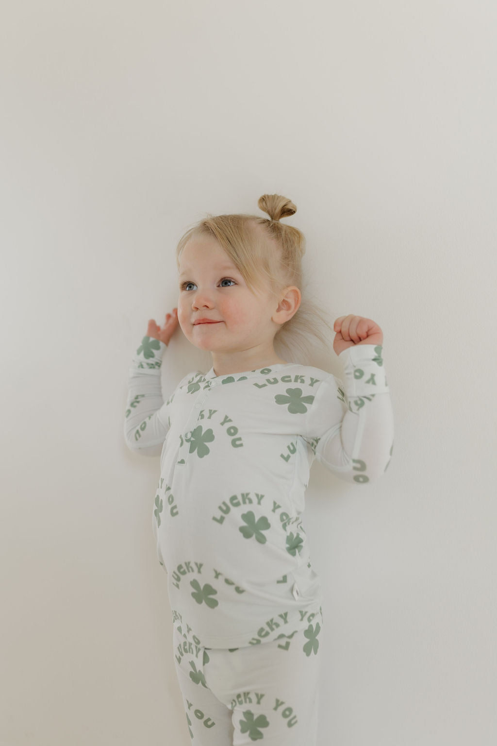 A young child with light hair in a topknot stands against a white wall, wearing forever french baby's Bamboo Two Piece Pajamas 'Lucky You' made of breathable, hypo-allergenic fabric. The pajamas feature green clovers as the child holds their hair up with both hands, smiling slightly to the side.