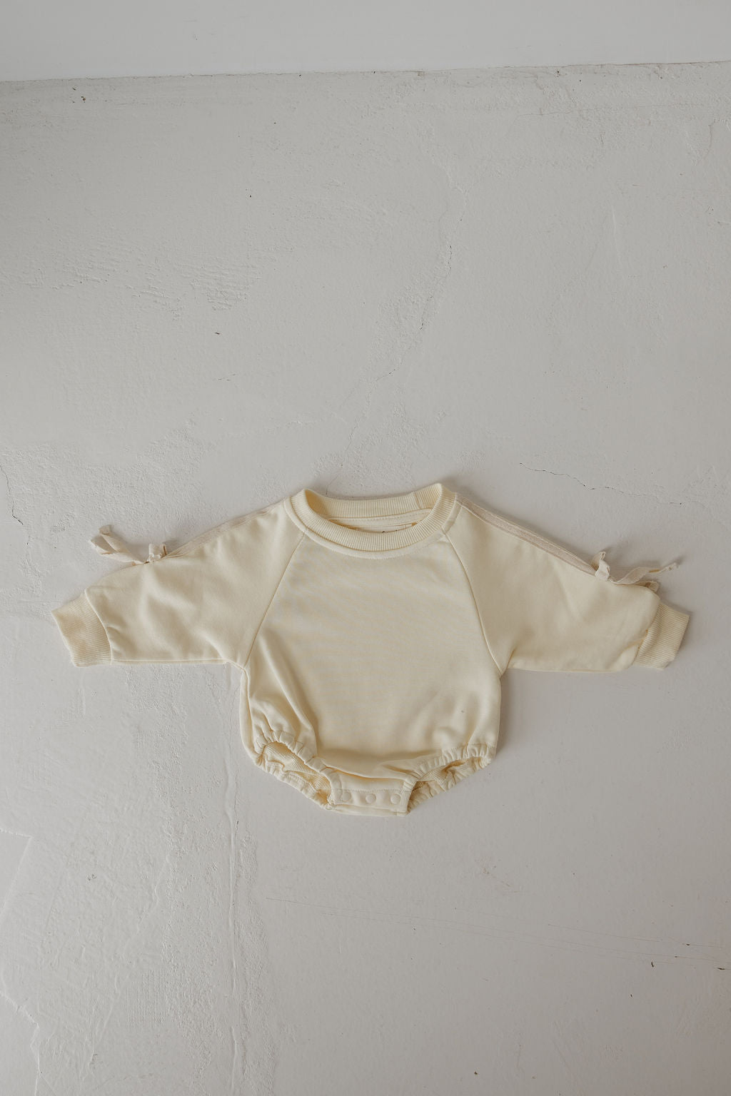 The forever french baby Sweatshirt Romper | Buttercream Bows is a cream, long-sleeve baby romper with side sleeve ties, elastic leg openings, and snap buttons—perfect for stylish newborns.