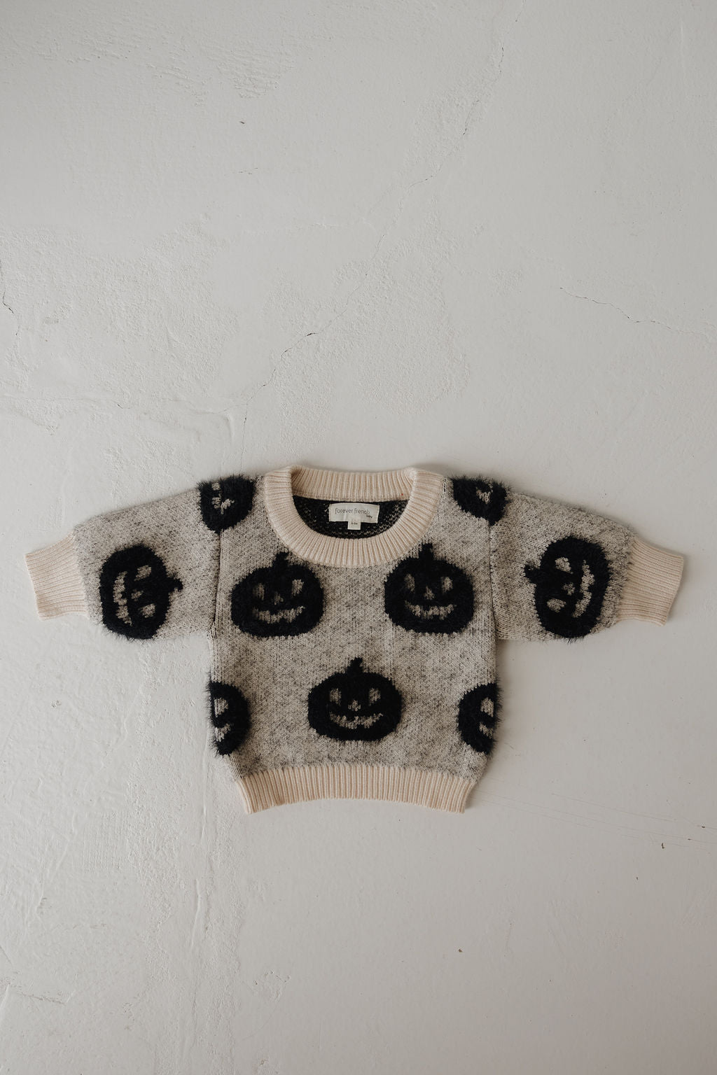 The Cozy Sweater | Halloween by forever french baby is a small, fuzzy cream sweater with long sleeves and black smiling jack-o'-lantern faces—an adorable Halloween essential, shown laid flat on a light textured surface.