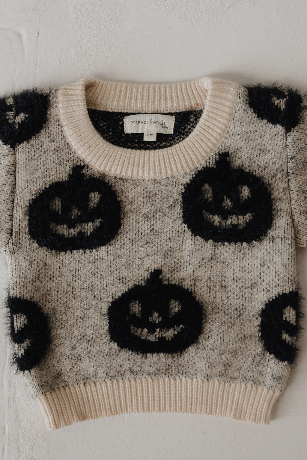 The forever french baby Cozy Sweater | Halloween is a light gray knit with a cream neckline and hem, featuring black fuzzy jack-o'-lantern faces. A cozy Halloween essential, it's shown laid flat on a white surface.
