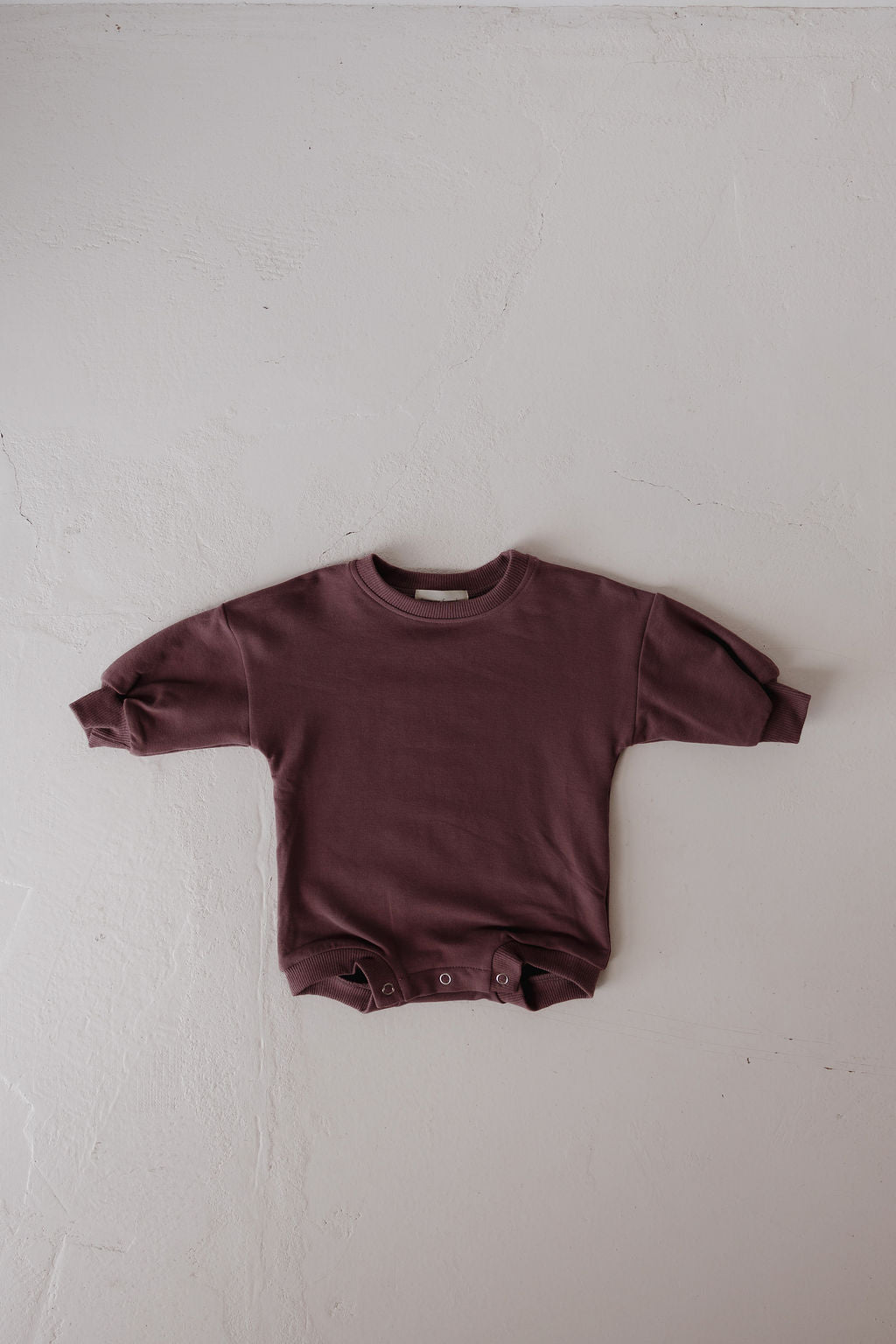 The forever french baby Sweatshirt Romper | Twilight, a long-sleeve maroon romper with bottom snap buttons, is laid flat on a light textured surface.