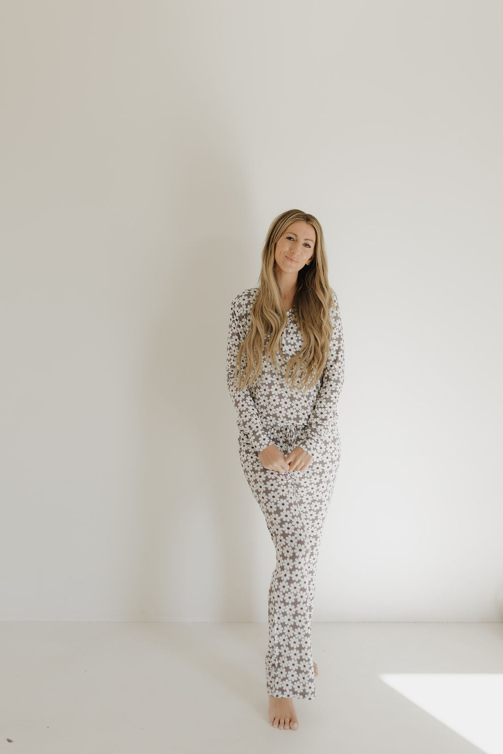 A person with long hair stands barefoot against a plain wall, wearing the floral-patterned Women's Bamboo Pajama by forever french baby. Their relaxed posture and gently clasped hands, combined with hypo-allergenic fabric, create a calm and cozy atmosphere.