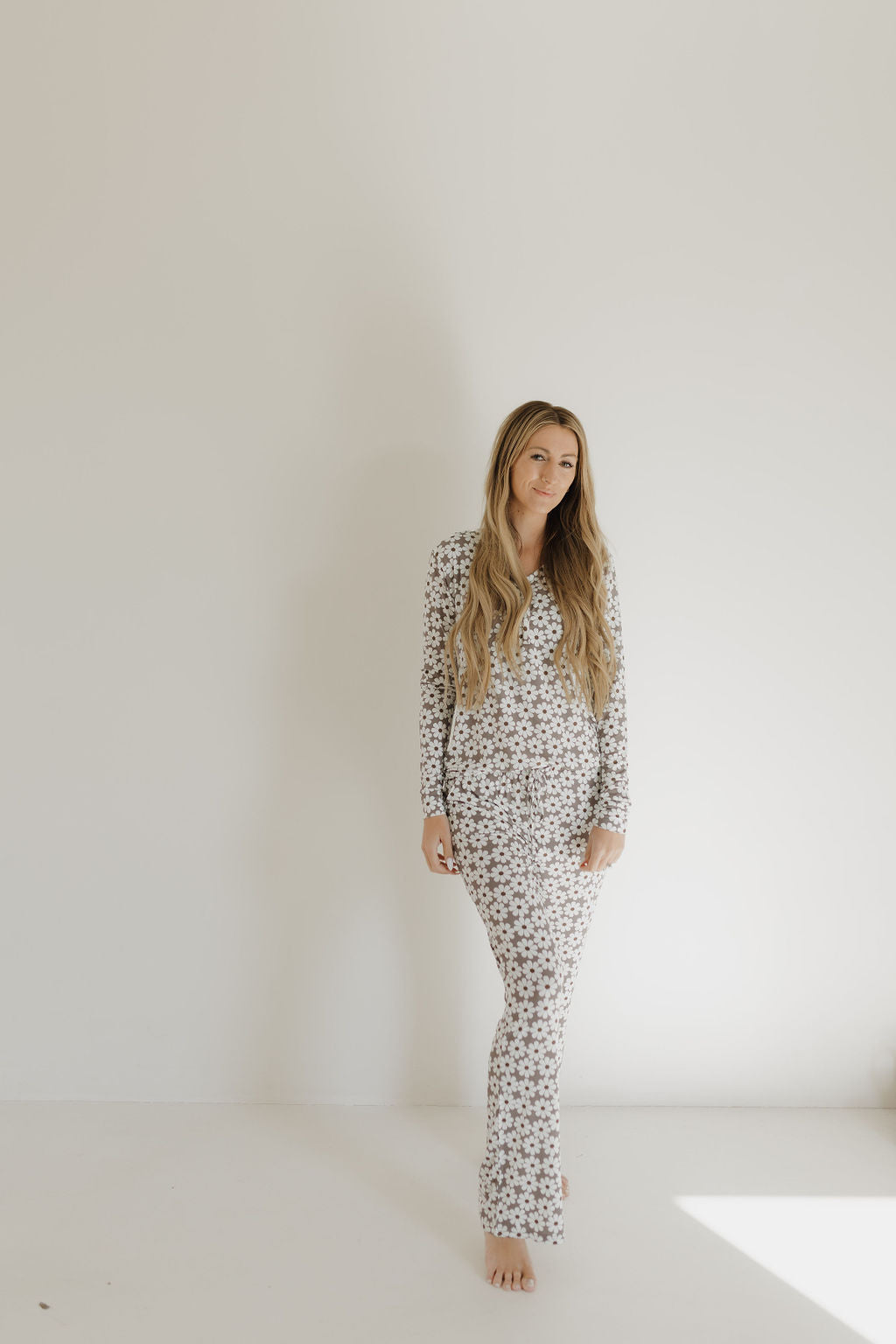 A person with long hair stands indoors against a plain light-colored wall, wearing a patterned long-sleeve top and matching Women's Bamboo Pajama by forever french baby. They look relaxed, enhanced by bright lighting that highlights the hypo-allergenic comfort of the Darlin' outfit.