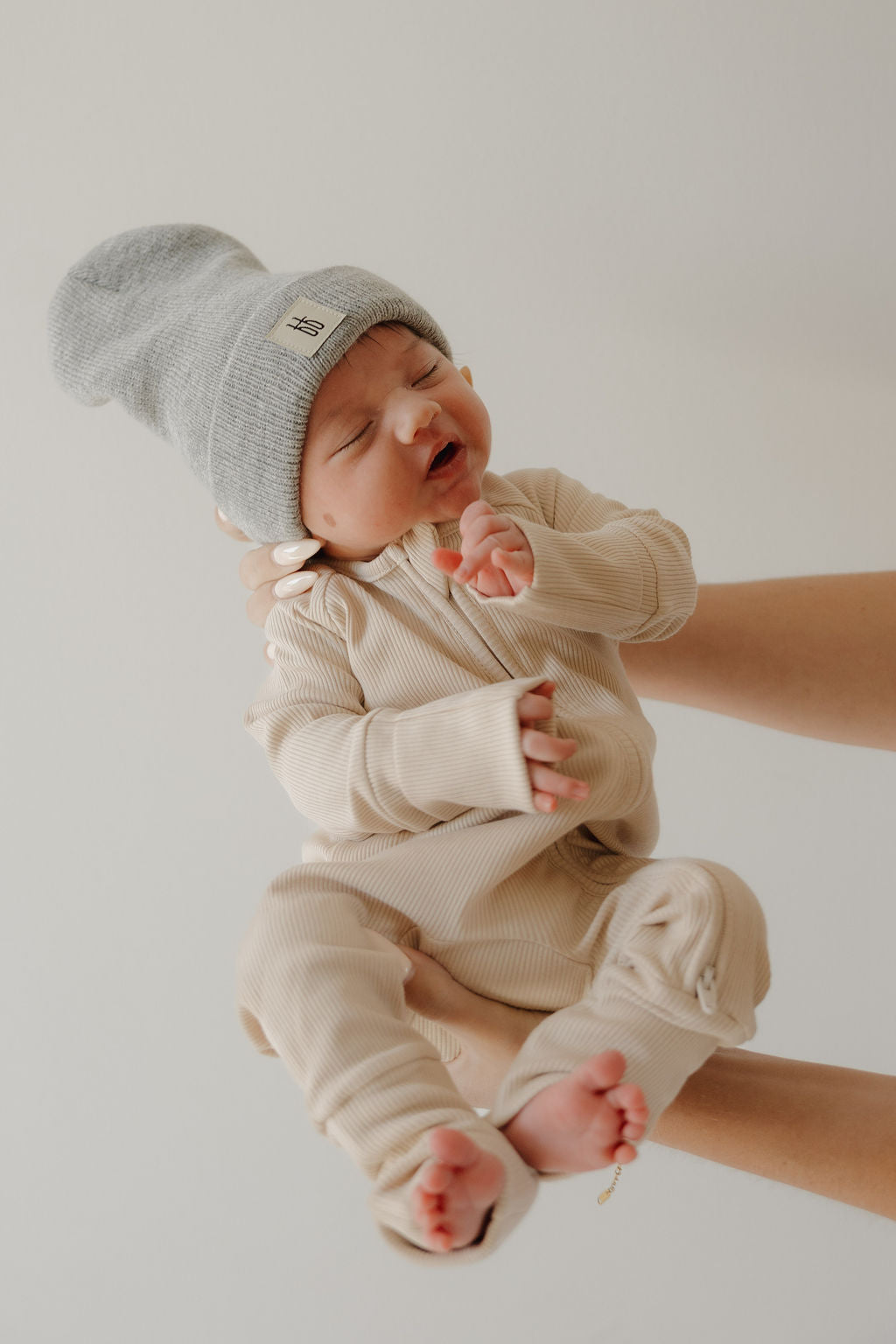 A baby wearing forever french baby's Bamboo Zip Pajamas | Ribbed Cream and a gray knit hat is gently held by an adult, face slightly scrunched as if about to cry, against a plain background.