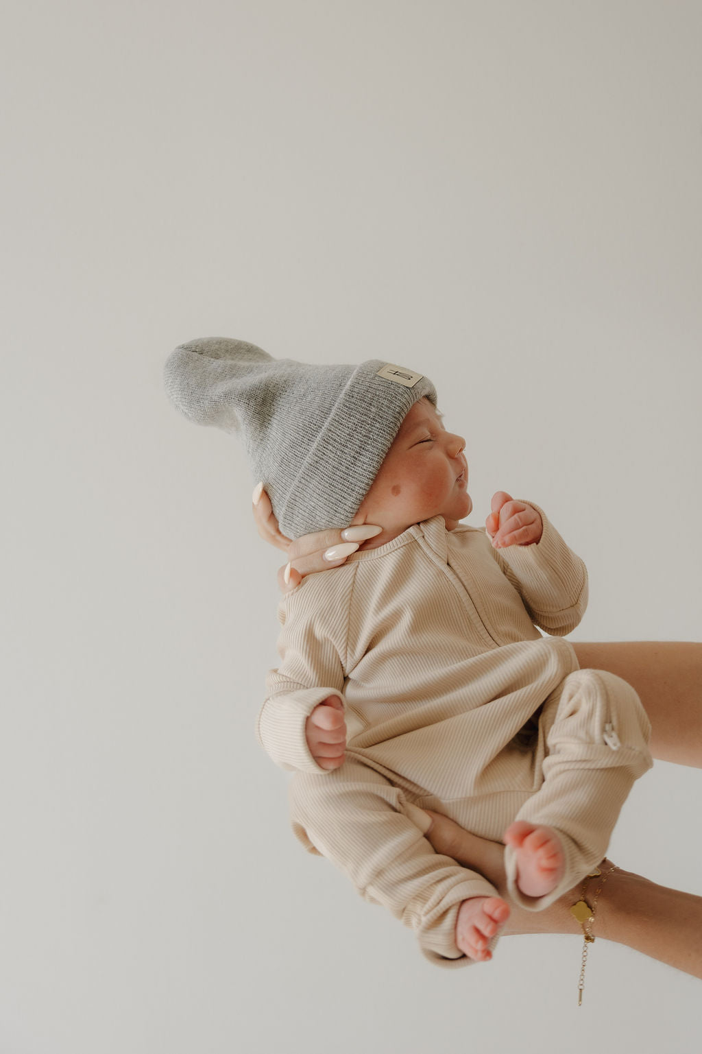 A newborn in forever french baby Bamboo Zip Pajamas | Ribbed Cream and a gray knit hat is gently held by an adult with manicured nails against a plain light background.