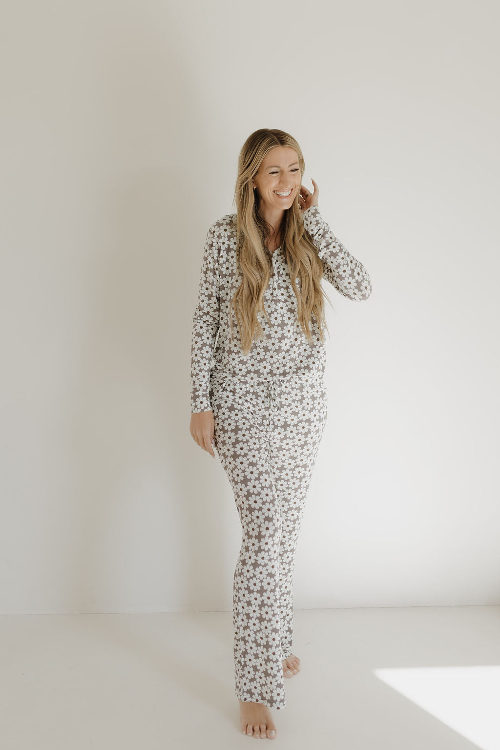 Long-haired person in a Women's Bamboo Pajama | Darlin' by forever french baby stands barefoot in a bright, minimalist room, smiling and posing with a hand near their ear.