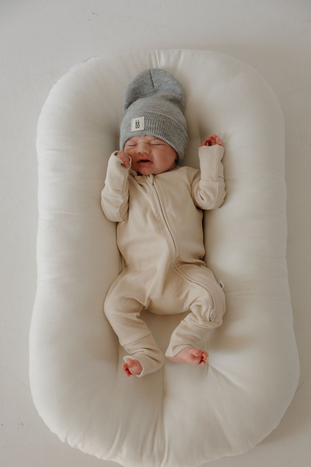 A newborn rests peacefully on a soft white pad, wearing forever french baby’s Bamboo Zip Pajamas in Ribbed Cream and a gray knit hat, with one hand near their face.