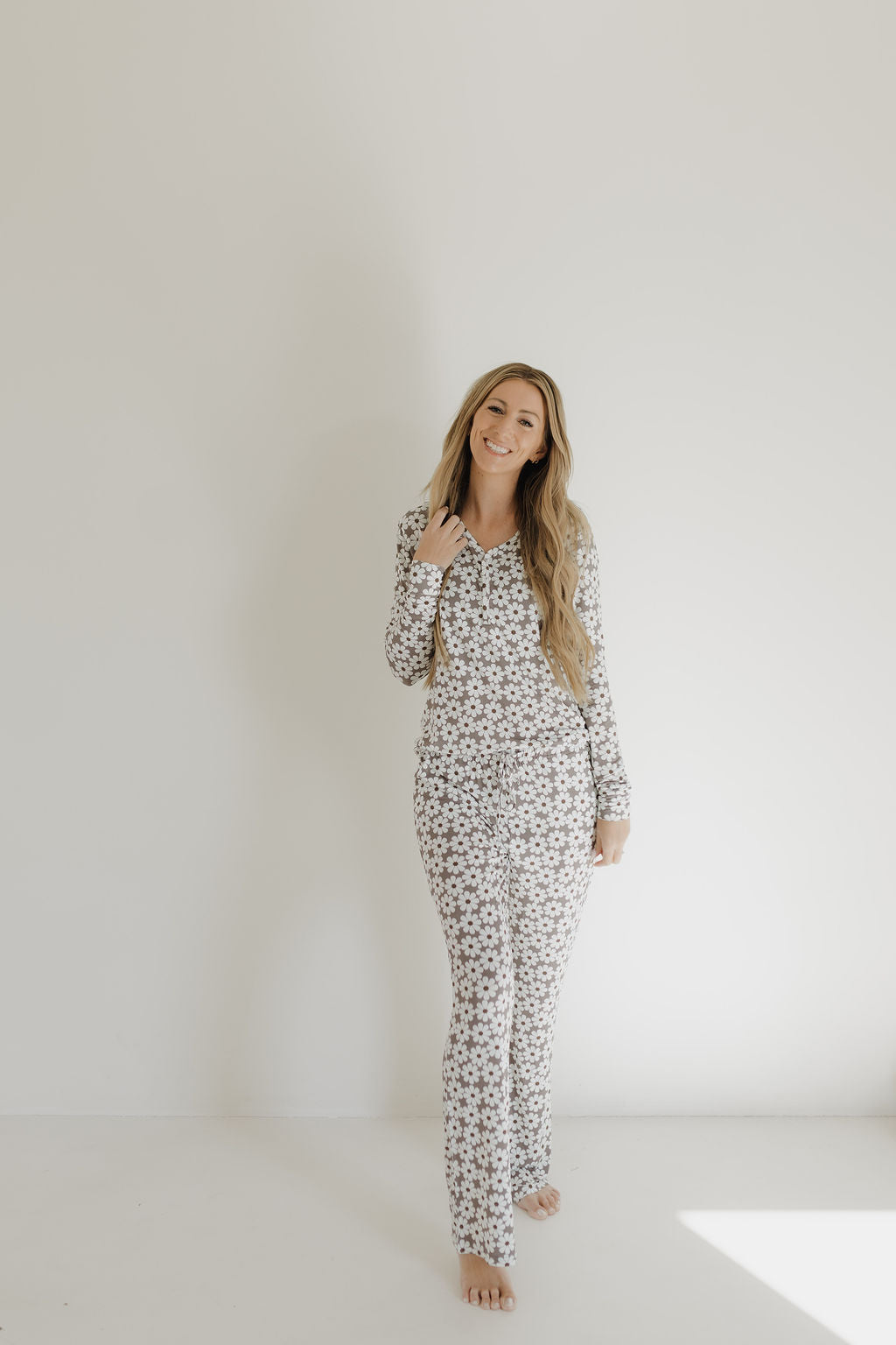 Standing barefoot against a white wall, a woman with long hair smiles as she holds her hair. She's wearing the 'Women's Bamboo Pajama | Darlin'' set from forever french baby, featuring a hypo-allergenic bamboo fabric with long sleeves and small floral patterns.