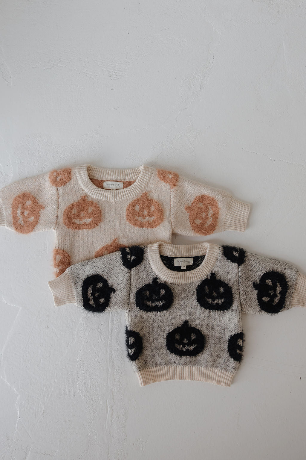 Two forever french baby Cozy Sweater | Halloween pieces are laid flat on a white surface—one cream with orange jack-o’-lanterns, the other gray with black—making them perfect Halloween essentials for kids.