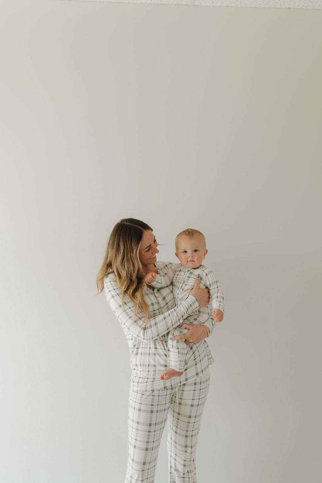 A woman cradles a baby, both wearing forever french baby's Bamboo Zip Pajamas in Picnic Plaid. Set against a light background, she smiles warmly at the baby, who curiously gazes towards the camera. The pajamas are made from breathable, hypo-allergenic bamboo fabric.