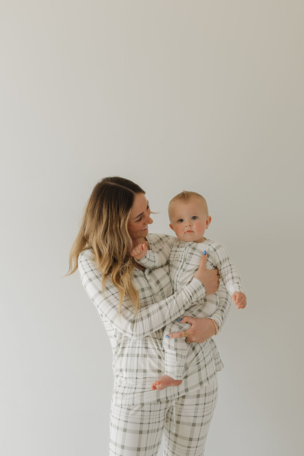 A woman smiles while holding a baby, both wearing forever french baby matching Picnic Plaid pajama sets crafted from breathable bamboo fabric. The light background highlights their hypoallergenic comfort as the baby gazes neutrally at the camera.