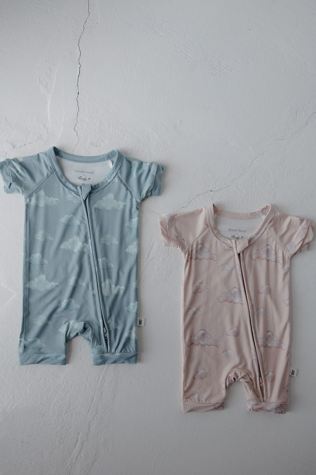 Two Kendy 3 Bamboo Zip Shortie | Duke rompers are on a white textured surface—one blue with cloud prints, one light pink with similar clouds. Both feature front zippers and offer breathable comfort for babies.