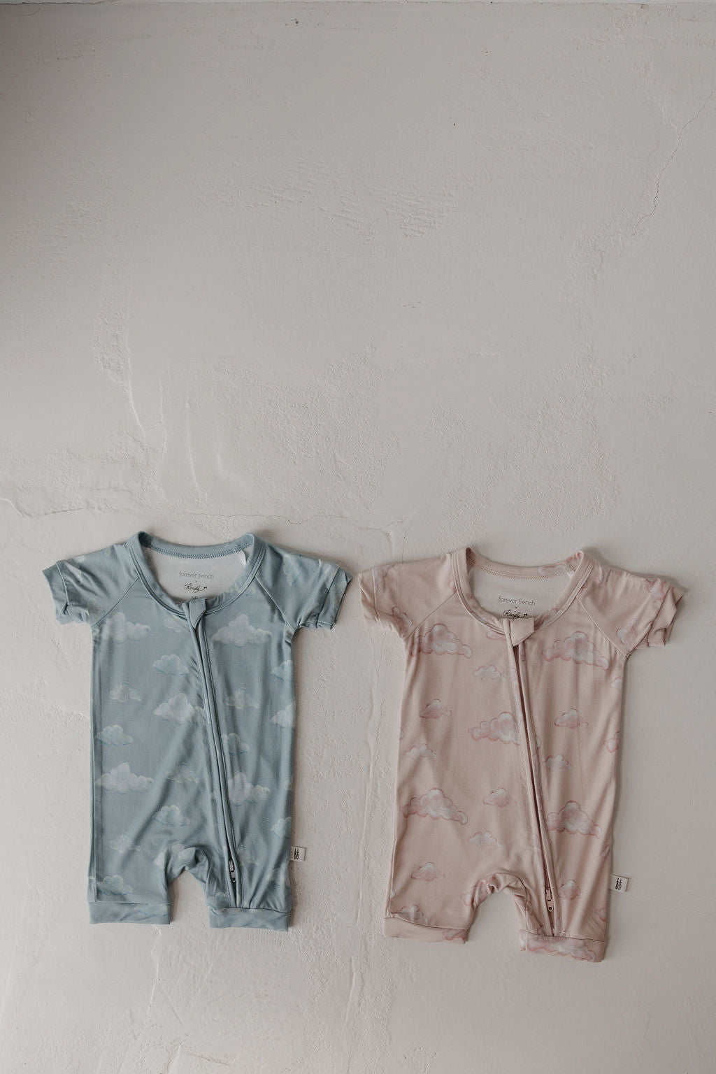 Two Kendy 3 Bamboo Zip Shortie rompers are laid flat: one blue with white clouds, the other pink with pink clouds. Both hypo-allergenic pieces have convenient front zippers and are breathable for baby comfort.