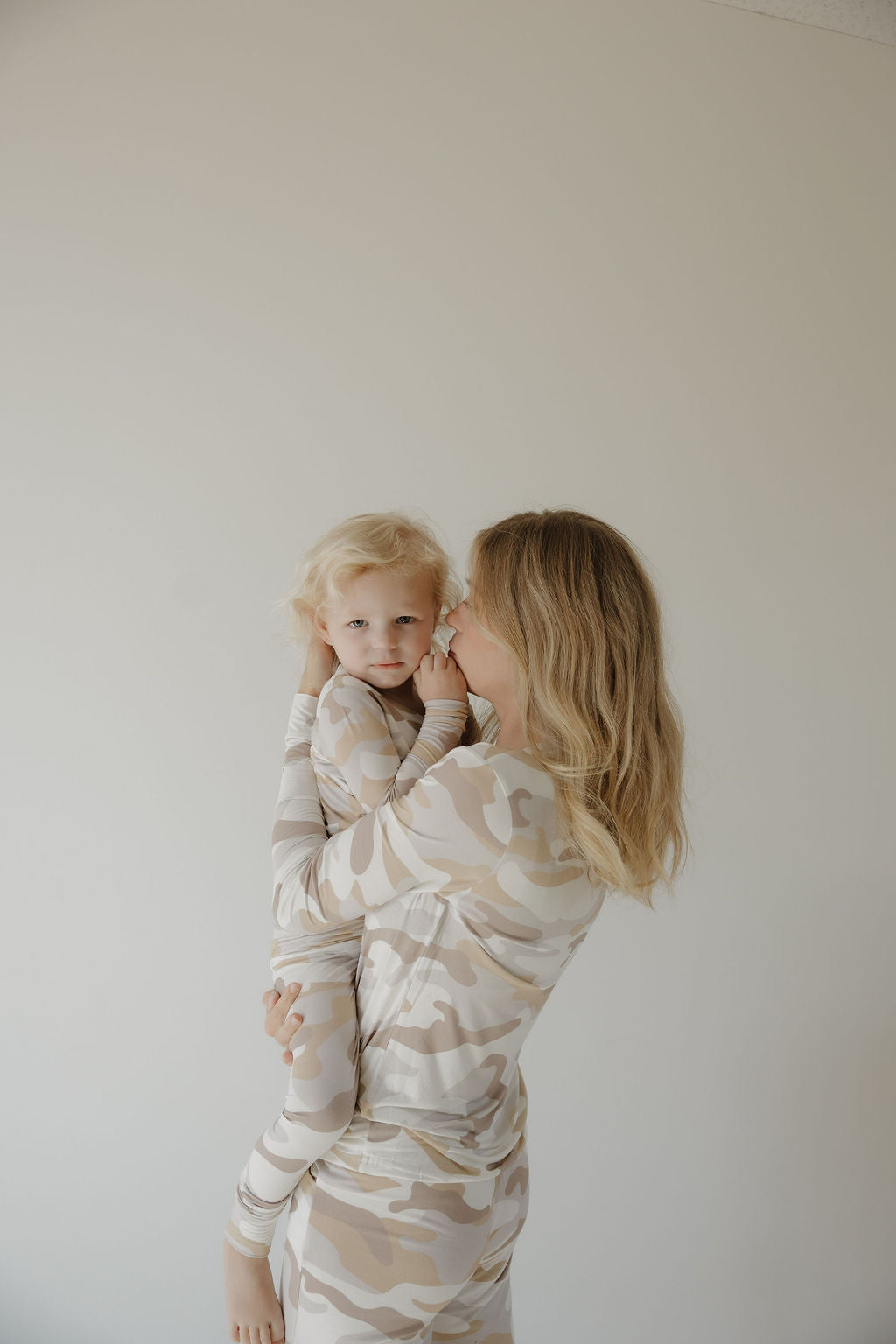 A woman with shoulder-length blonde hair holds a young child, both wearing matching forever french baby Bamboo Two Piece Pajamas | Camo 2.0, known for being hypo-allergenic, against a plain light background.