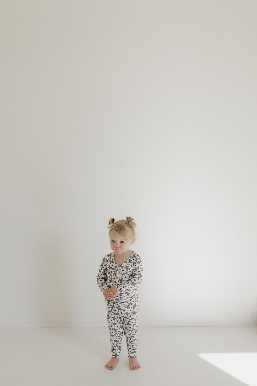 A young child with light hair styled in two small buns stands barefoot against a plain white background, wearing the "Bamboo Two Piece Pajamas | Darlin'" by forever french baby. This long-sleeved, floral-patterned hypo-allergenic outfit is made from bamboo clothing, and the child gazes slightly to the left.